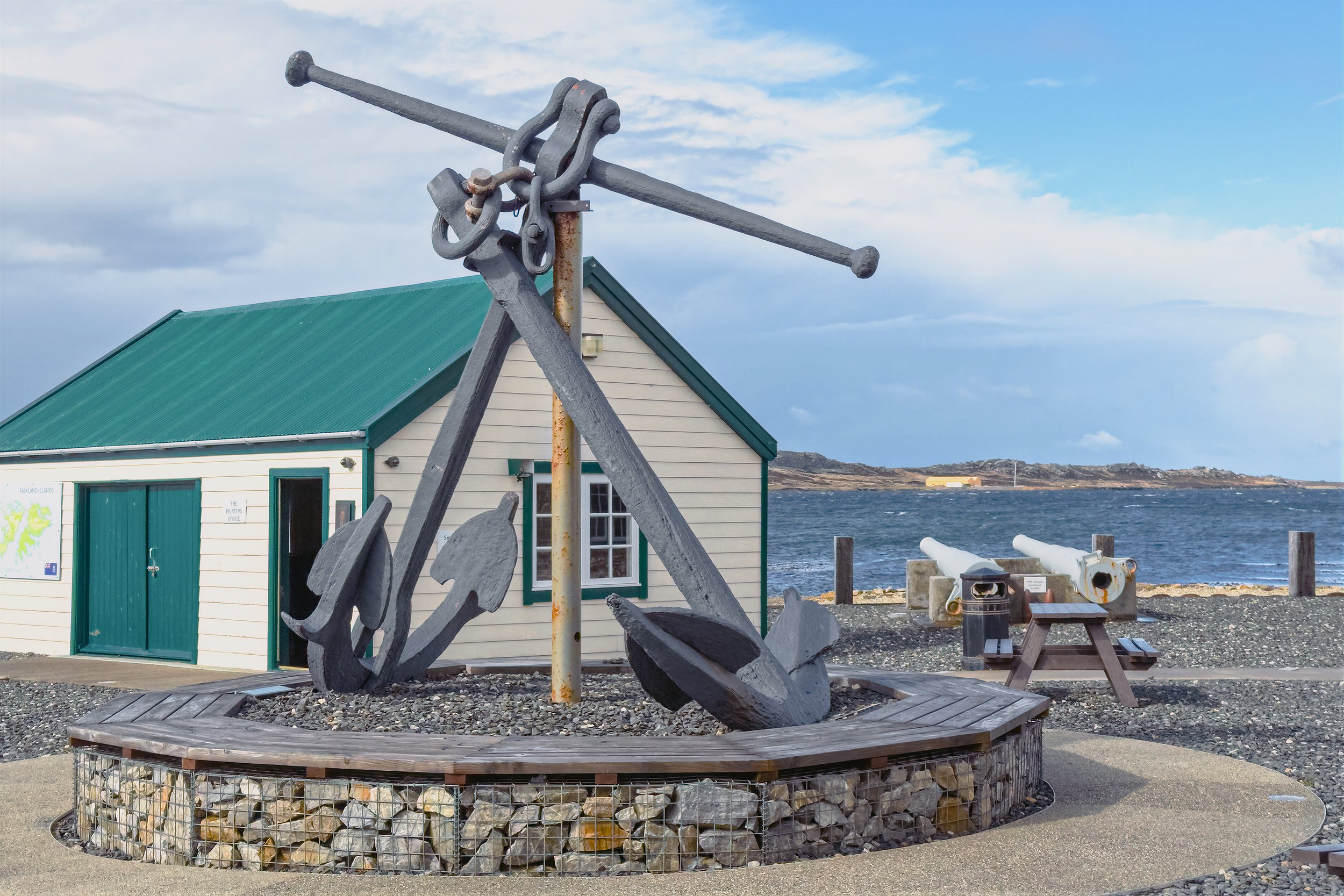 A large gray anchor sculpture is displayed in front of a building with a green roof and white walls by the water, with a cloudy sky overhead.