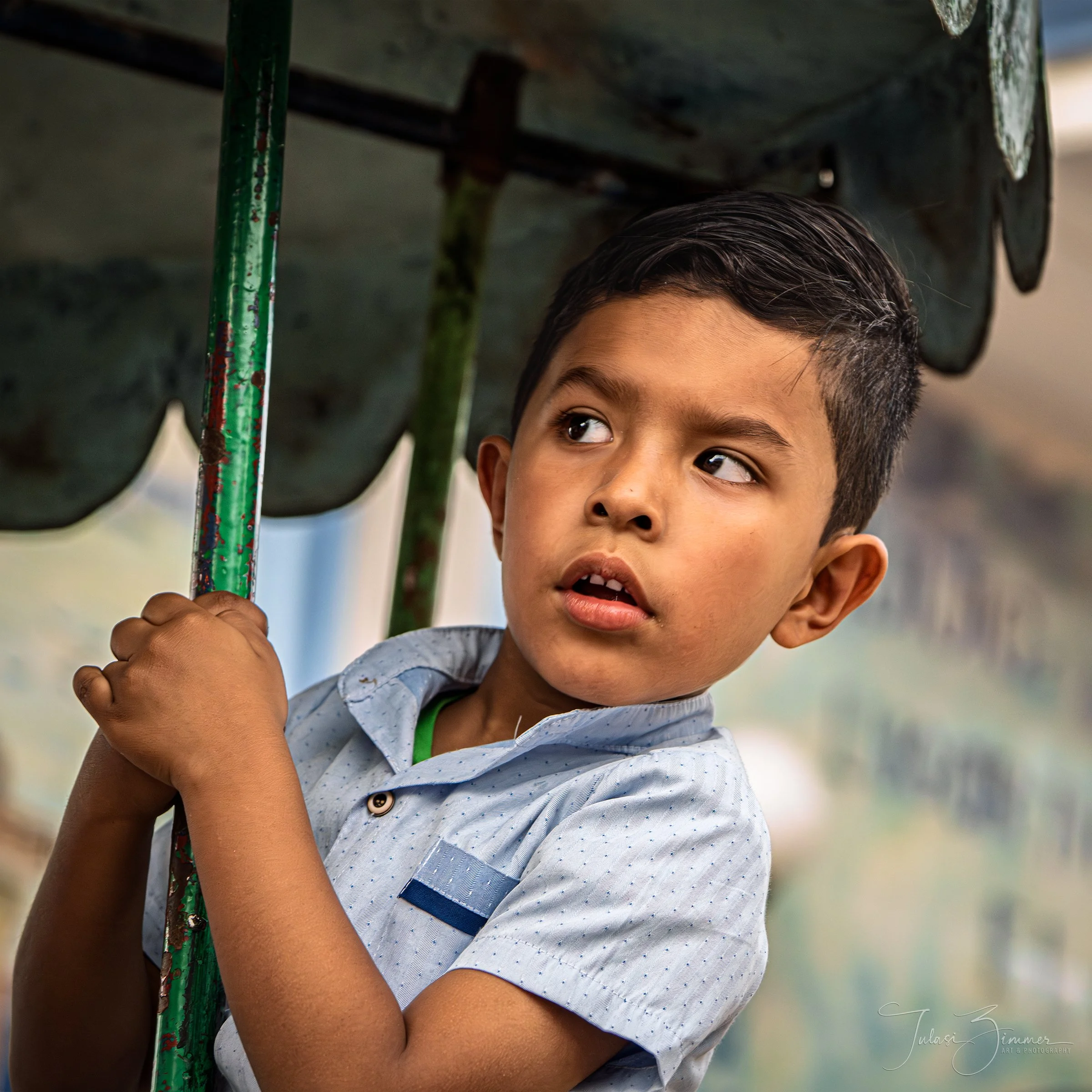 Close-up of a young Mexican boy with short dark hair and brown eyes, holding a green pole with red paint splatters, looking over his shoulder outdoors.