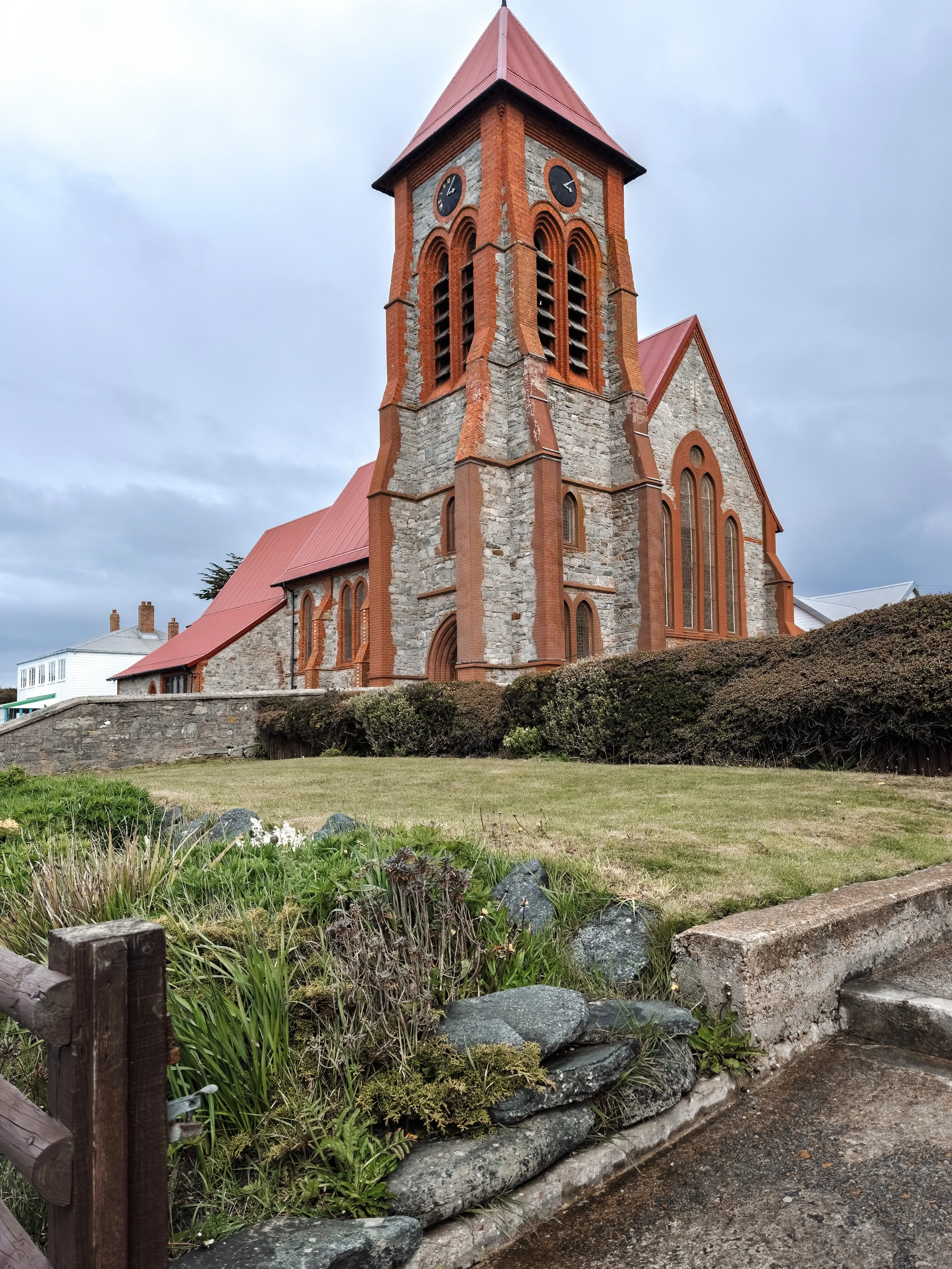 A stone church with red brick accents and a red roof, featuring a tall clock tower with two clocks, set against a cloudy sky and grassy foreground with rocks and plants.