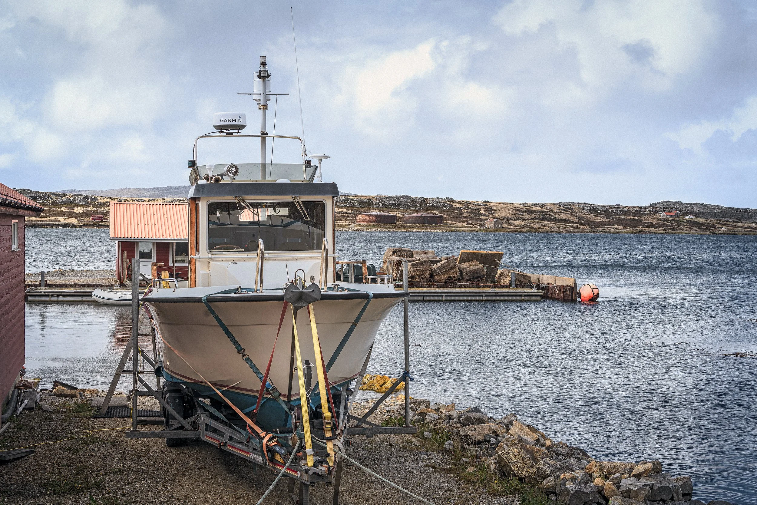 A boat is on a trailer on a rocky shoreline near water, with buildings and hills in the background under a cloudy sky.