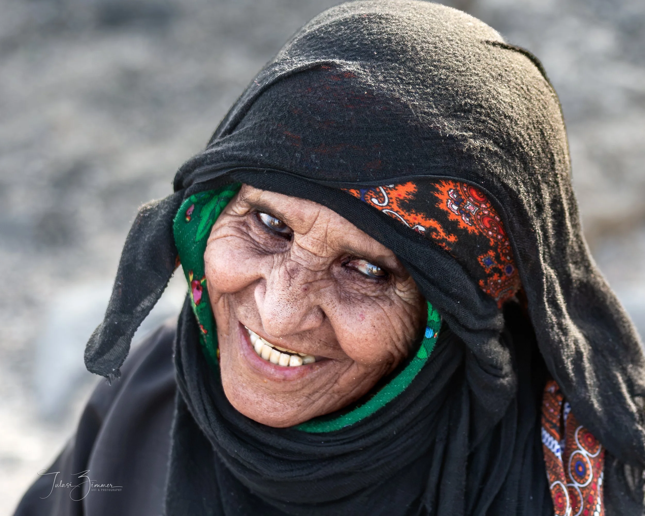 Close-up of an elderly woman smiling, wearing a black headscarf with colorful patterns and minor dust or dirt on her head covering.