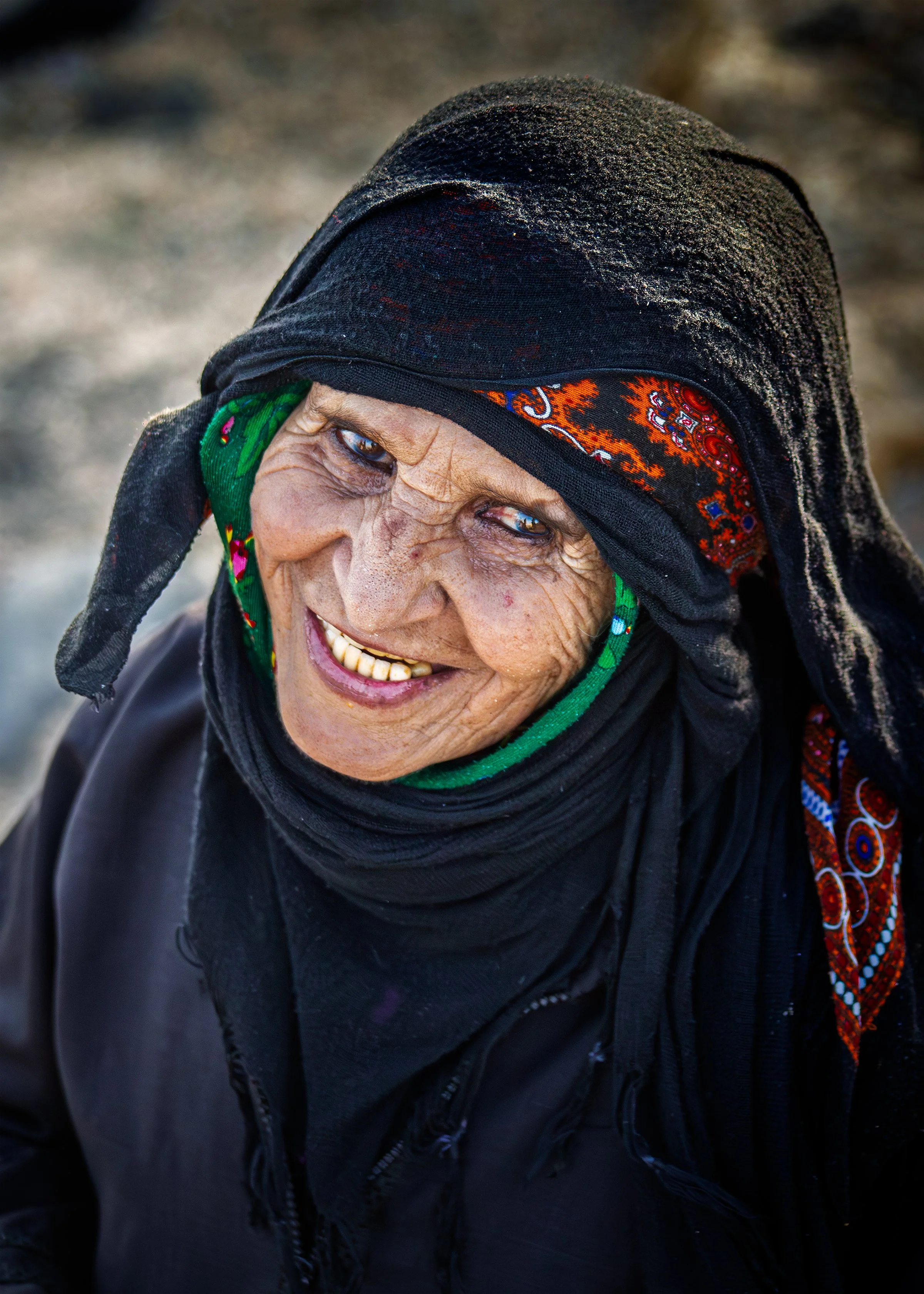 Close-up of an elderly Saudi woman wearing a black headscarf with colorful patterns, smiling widely, with a cheerful expression and deep wrinkles on her face.