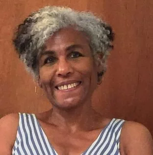 Smiling woman with short gray and black hair wearing a blue and white striped sleeveless top, against a wooden background.