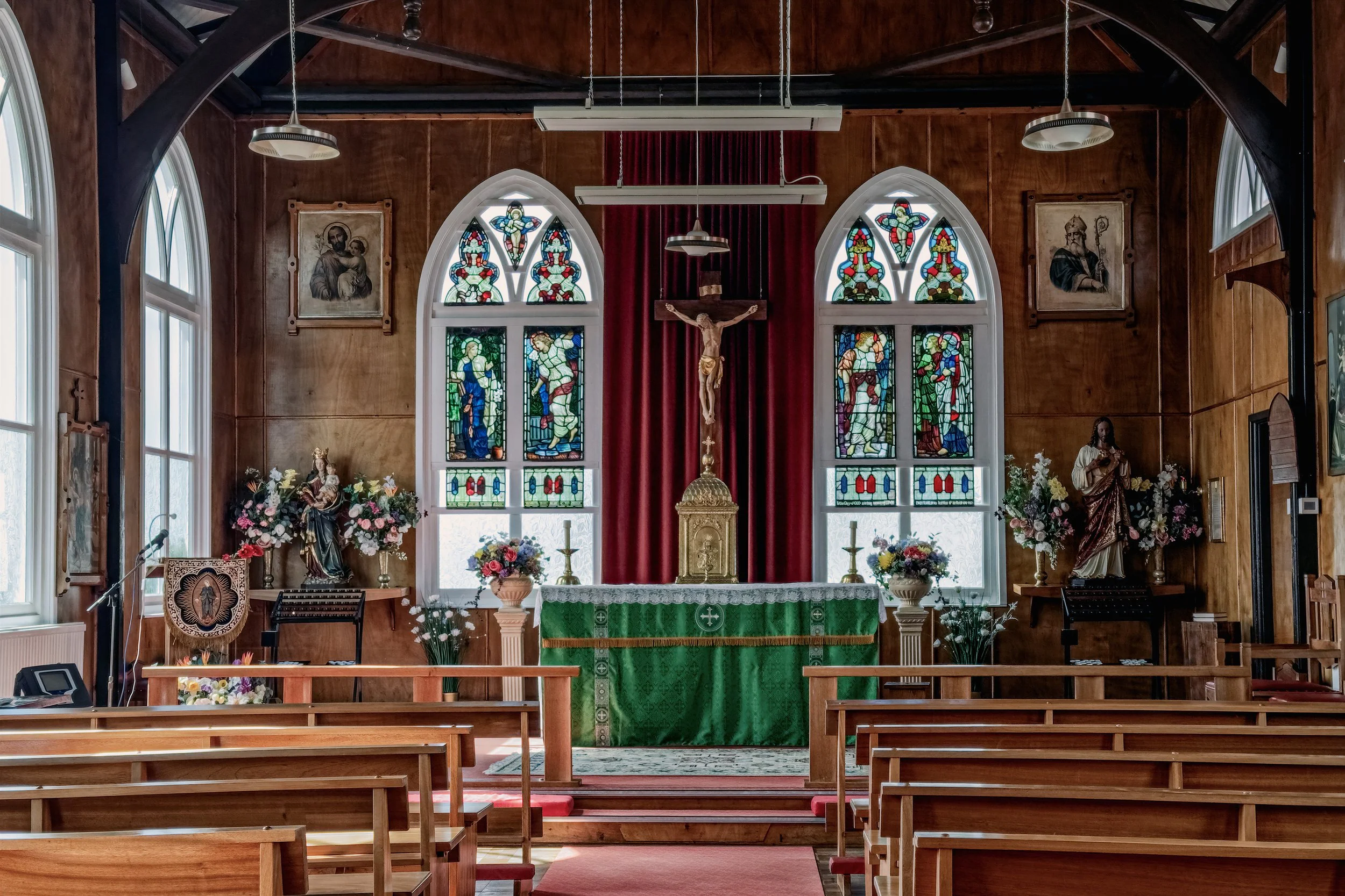 Interior of a church with stained glass windows, wooden walls, altar, crucifix, statues, and floral arrangements.