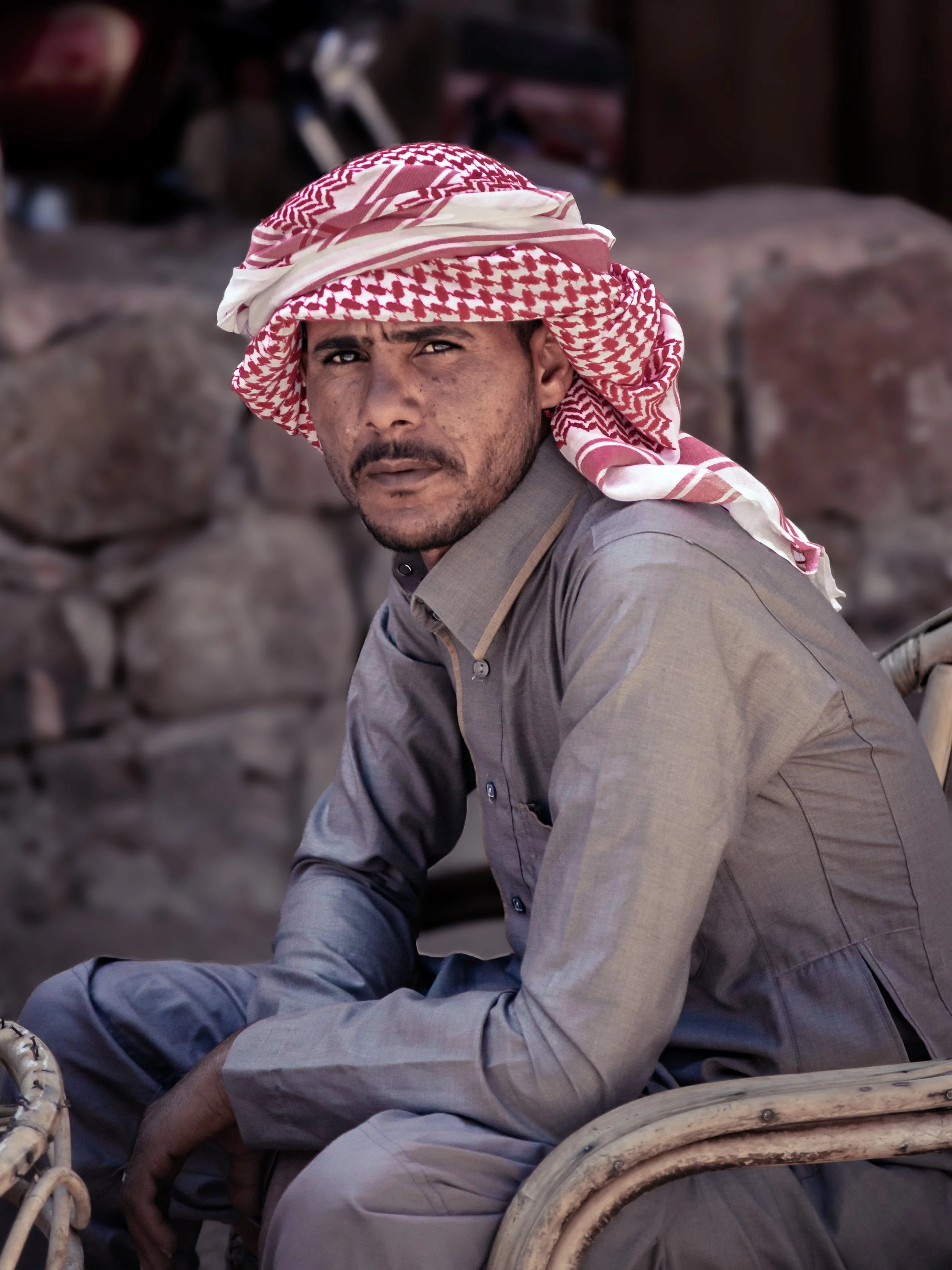 A man wearing a red and white keffiyeh sitting on a traditional woven chair, with a focused expression, in an outdoor setting with rocks and blurred objects in the background.