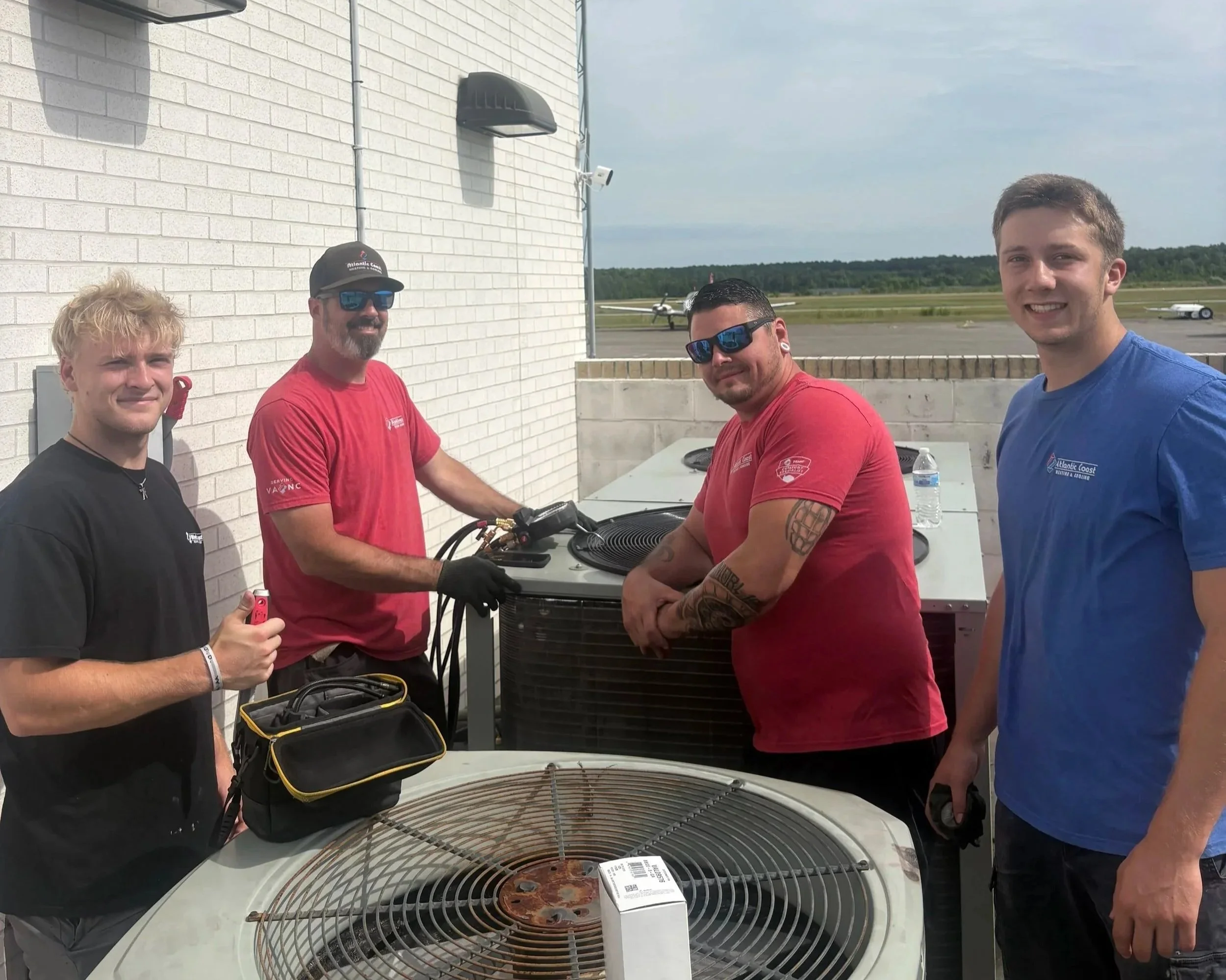 Four men stand around a table with a grill, preparing to cook pizza outdoors. They are smiling, wearing casual shirts and sunglasses, and appear to be on a rooftop or outdoor patio with a view of an airport runway in the background.