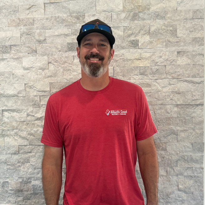 A man with a beard wearing a red t-shirt and a baseball cap standing in front of a gray brick wall.