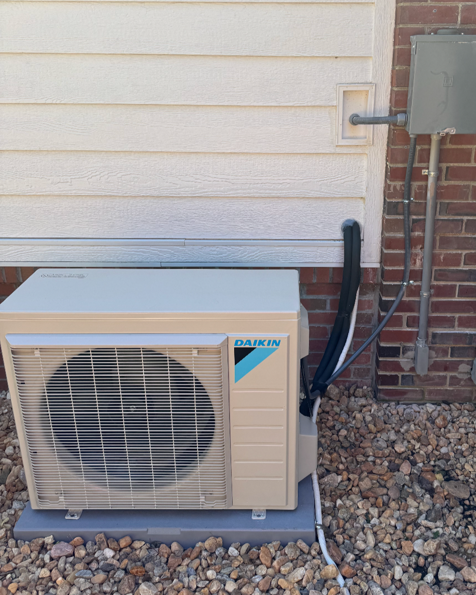 Outdoor air conditioning unit installed next to a house wall with gravel ground and electrical wiring.