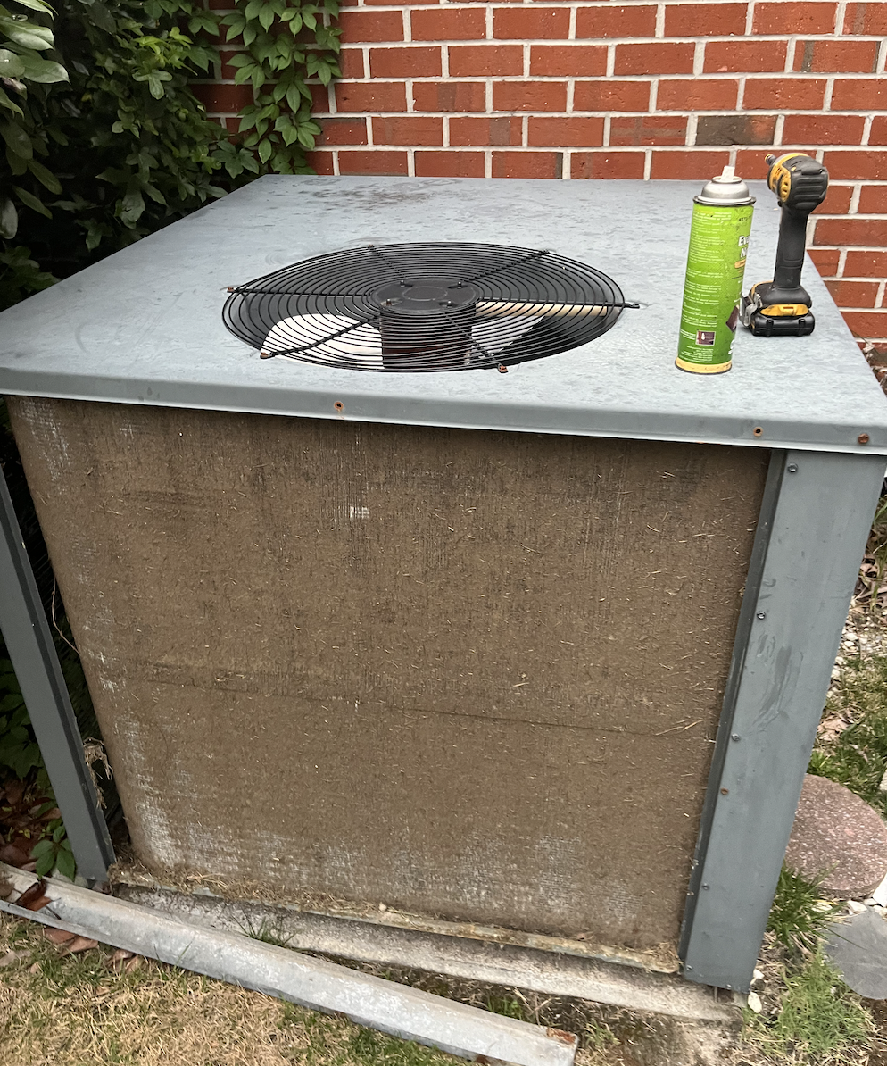 An outdoor air conditioning unit with a fan, partially covered with a protective grill, mounted on a concrete slab next to a brick wall. Tools and spray can are on top of the unit.