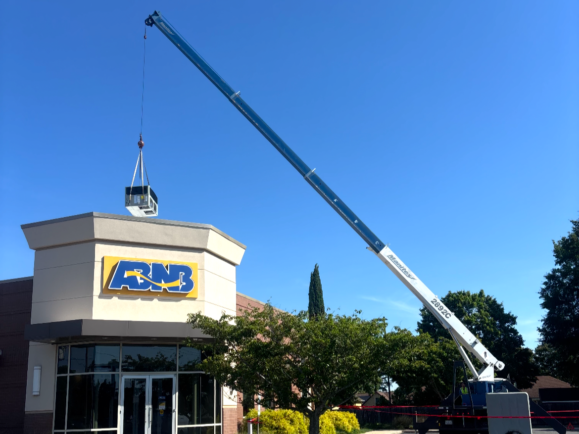 A crane lifting a large object above the ABNB building with a clear blue sky and trees in the background.