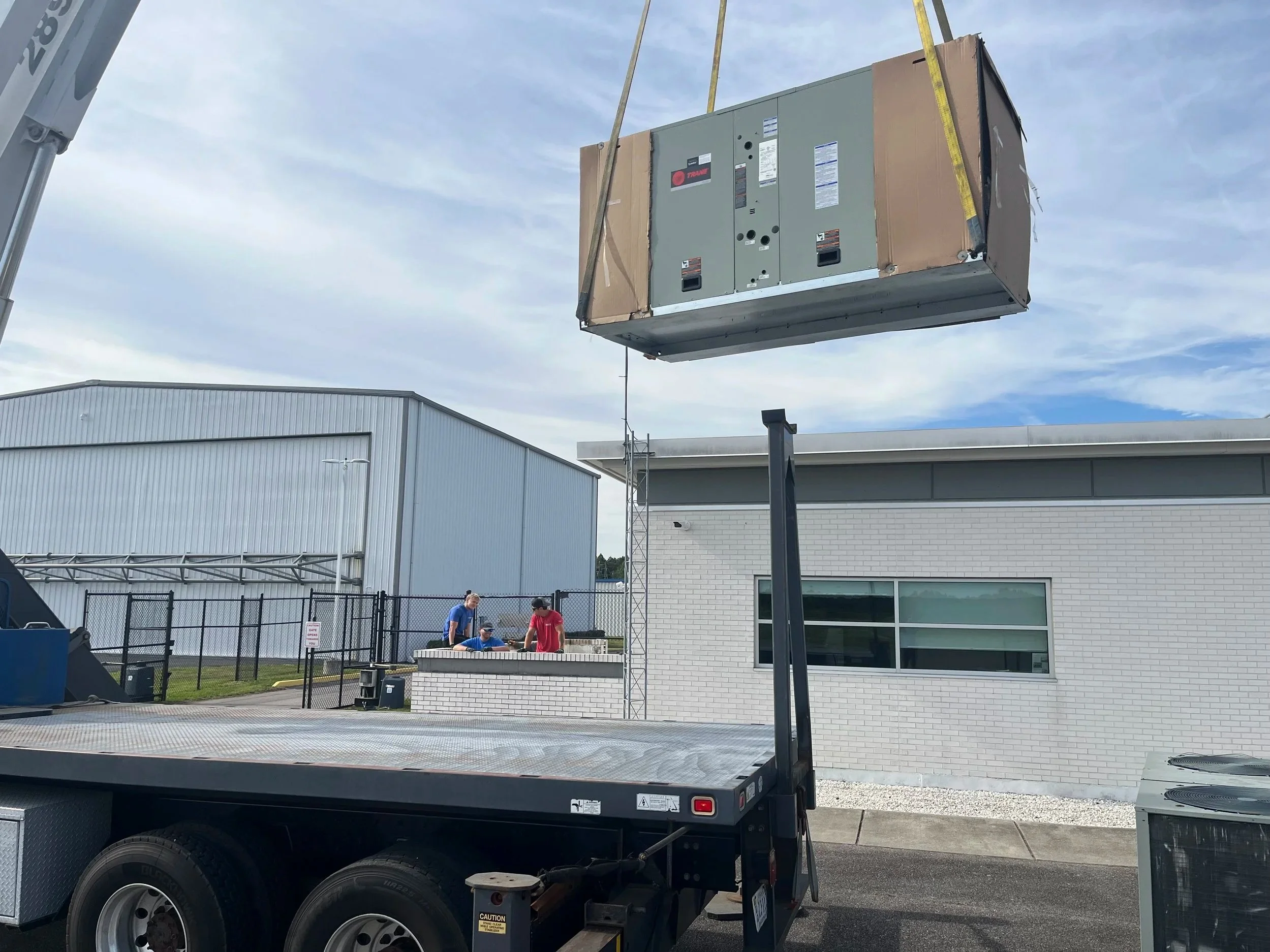 A crane lifting a large air conditioning unit or HVAC equipment onto a building or roof. The building has a gray exterior with metal siding and a white brick facade. There are three people on the ground observing or assisting with the installation, a