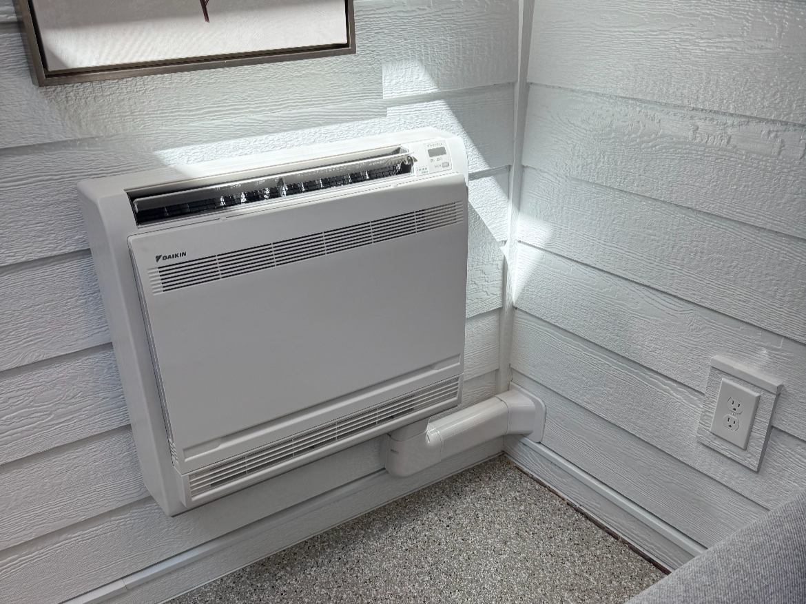 A wall-mounted Daikin electric heater with a vent below, near a corner with textured white siding and an electrical outlet.