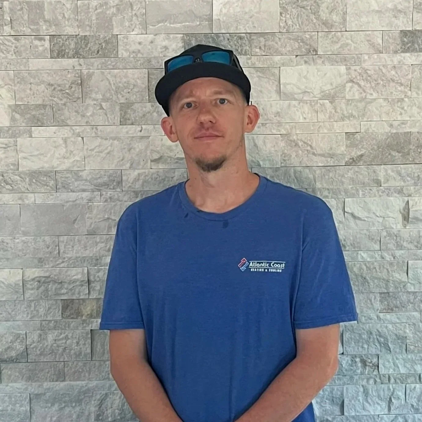 A man wearing a black cap with sunglasses on top, a blue T-shirt from Atlantic Coast Heating & Cooling, standing in front of a gray stone wall.