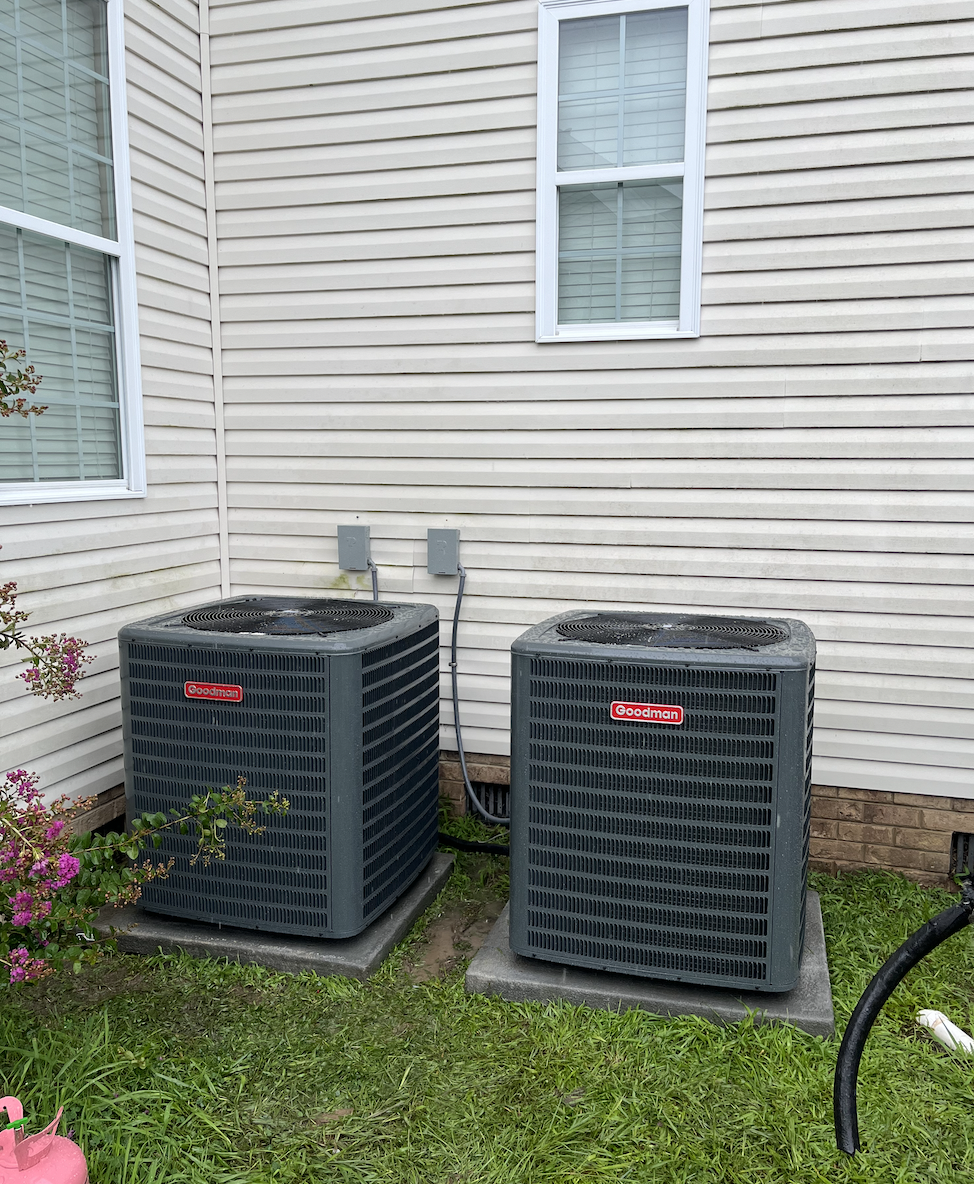 Two Goodman air conditioning units outside a house with beige vinyl siding, positioned on cement pads, surrounded by grass and bushes.