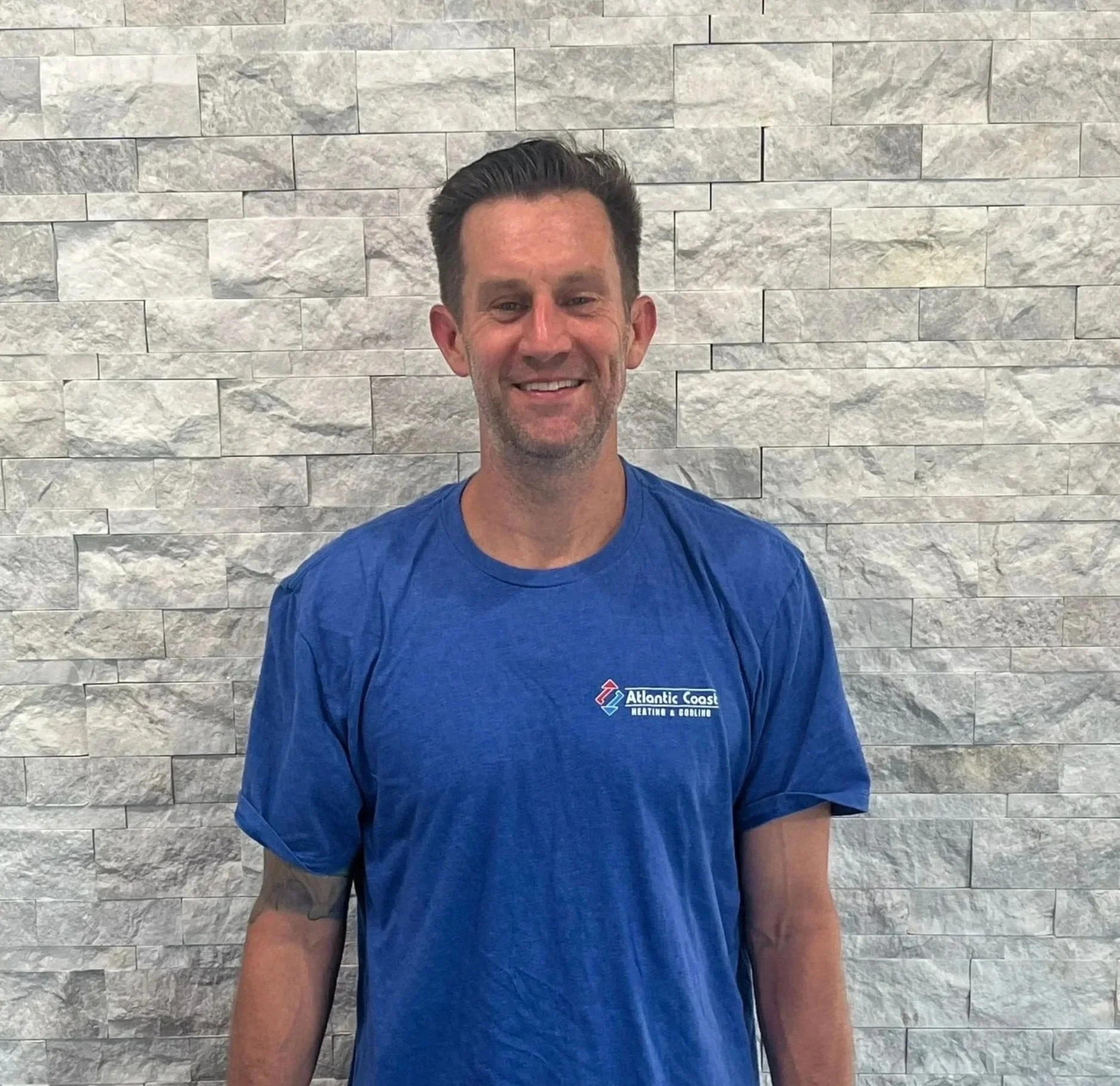 A man with short dark hair and a broad smile, wearing a blue T-shirt with a logo that reads 'Atlantic Coast Heating & Cooling,' standing in front of a textured white brick wall.