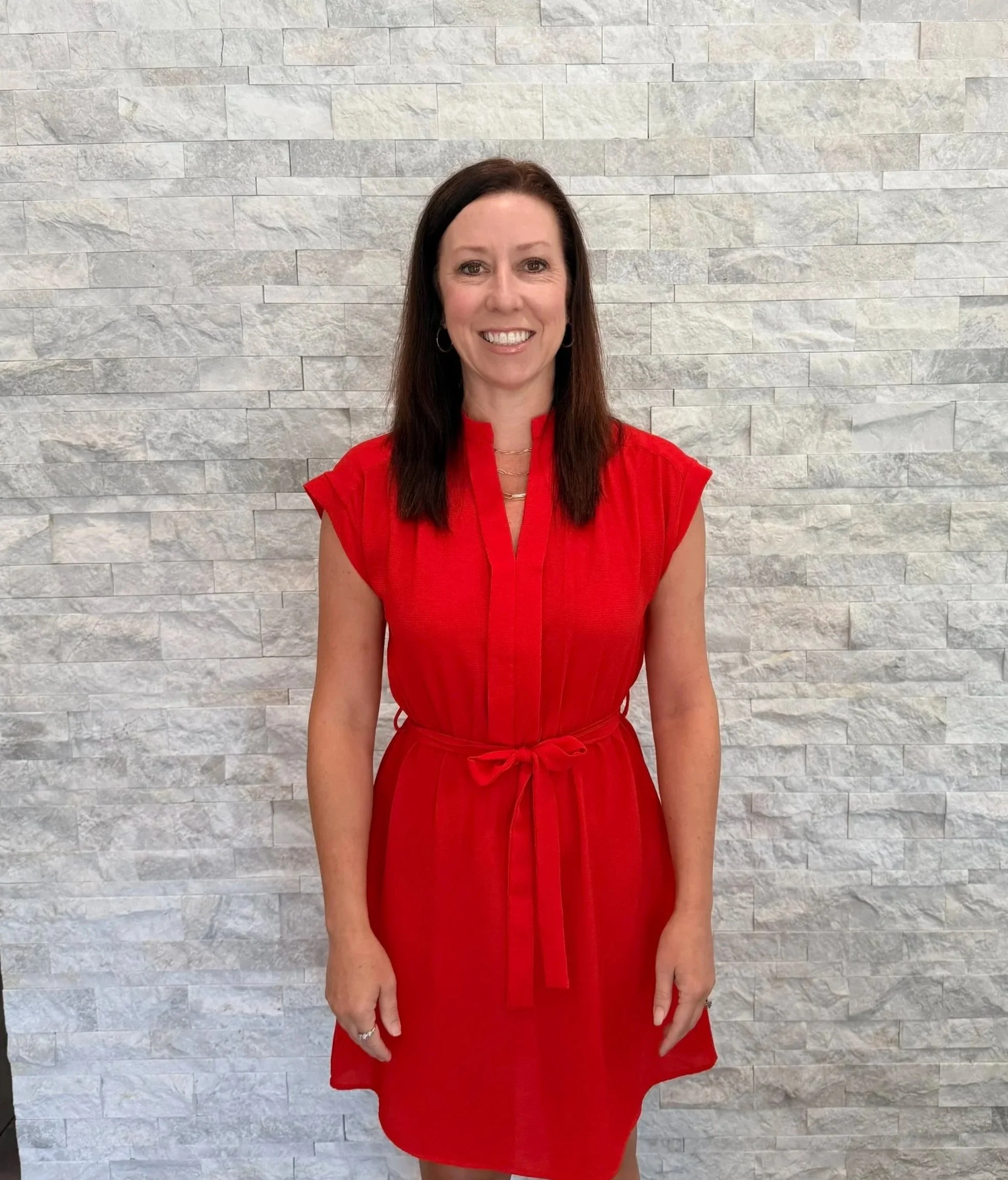 A woman with shoulder-length dark brown hair and hoop earrings, wearing a red dress, smiling, standing in front of a light-colored stone wall.