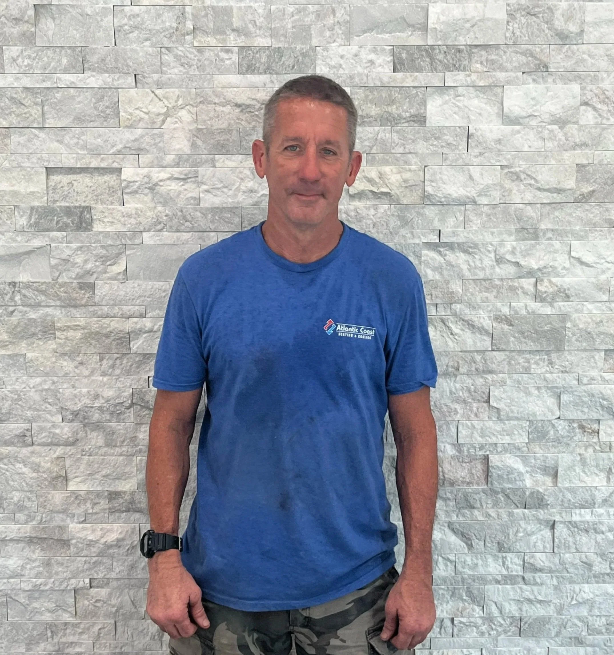 A middle-aged man wearing a blue T-shirt and camouflage shorts standing in front of a gray stone wall.