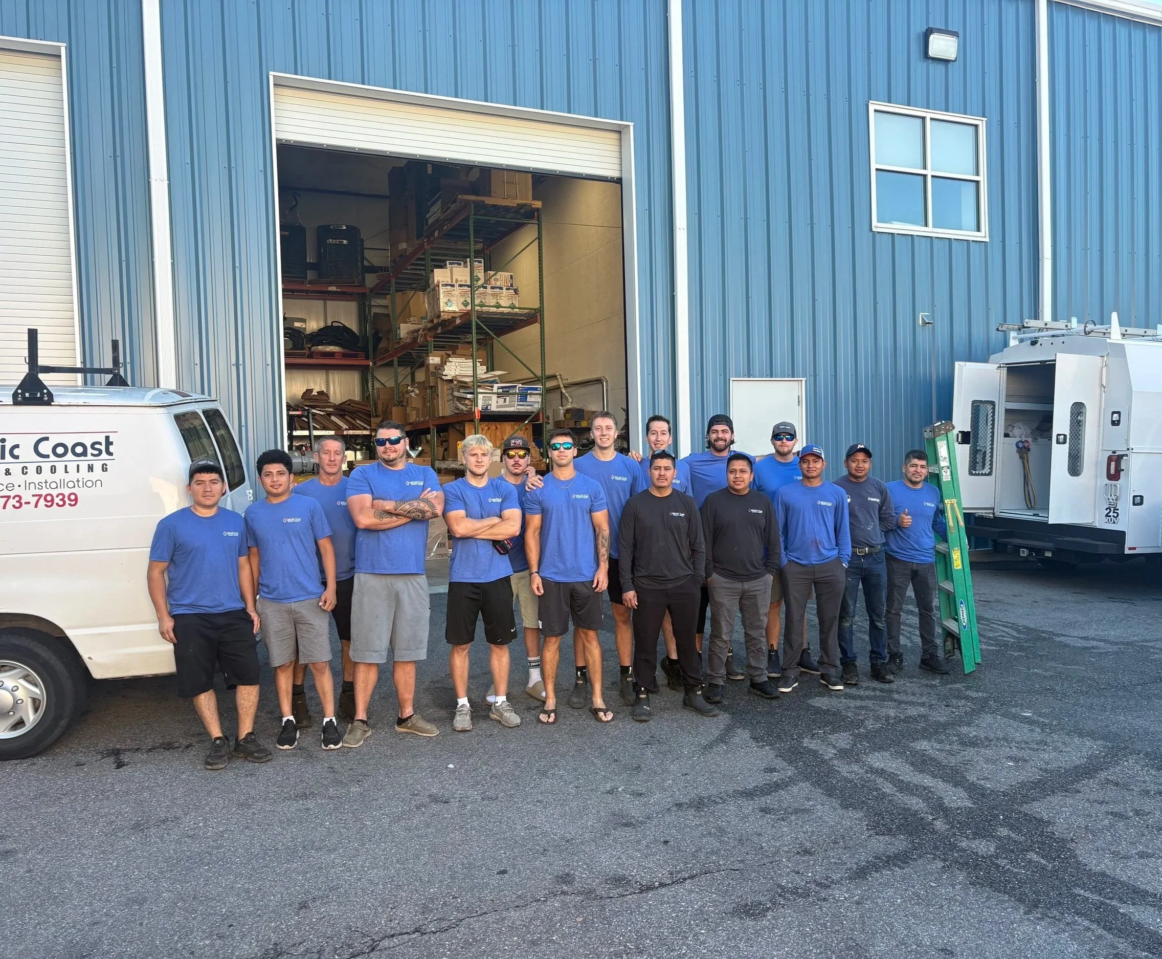 Group of workers standing in front of a blue industrial building with open garage door filled with storage shelves and equipment.