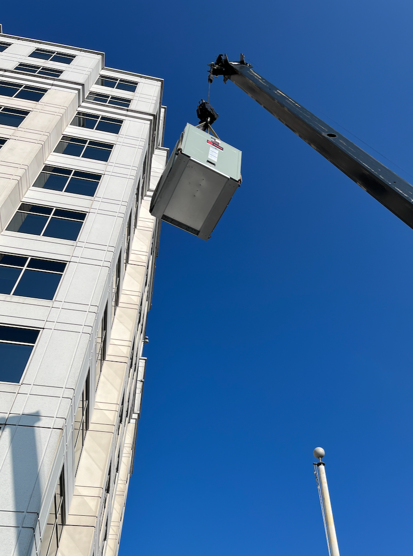 A construction crane lifting a large electrical or utility box beside a tall, white office building with multiple windows, against a clear blue sky.