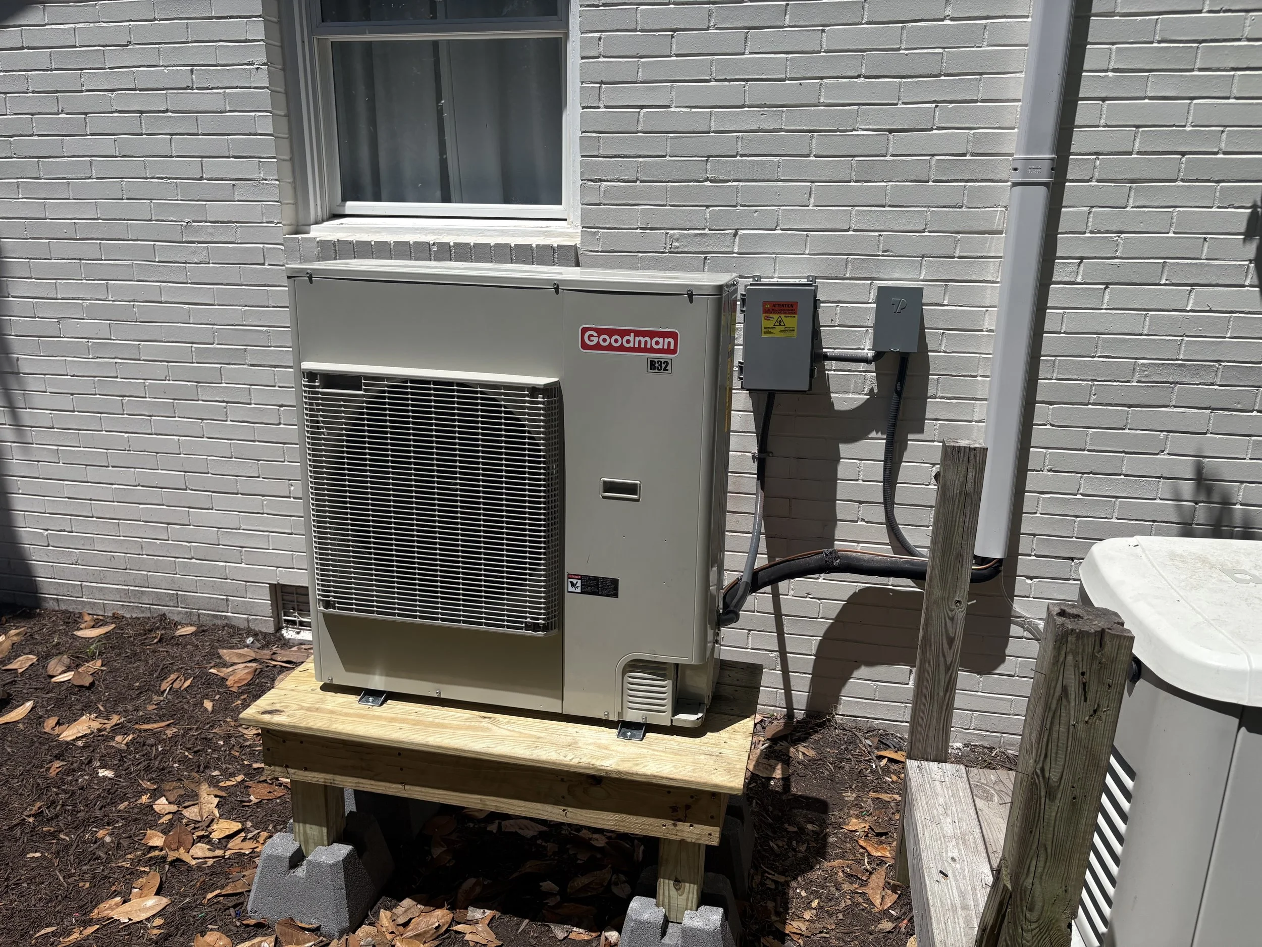 An outdoor air conditioning unit is placed on a wooden platform against a white brick wall. The unit has a