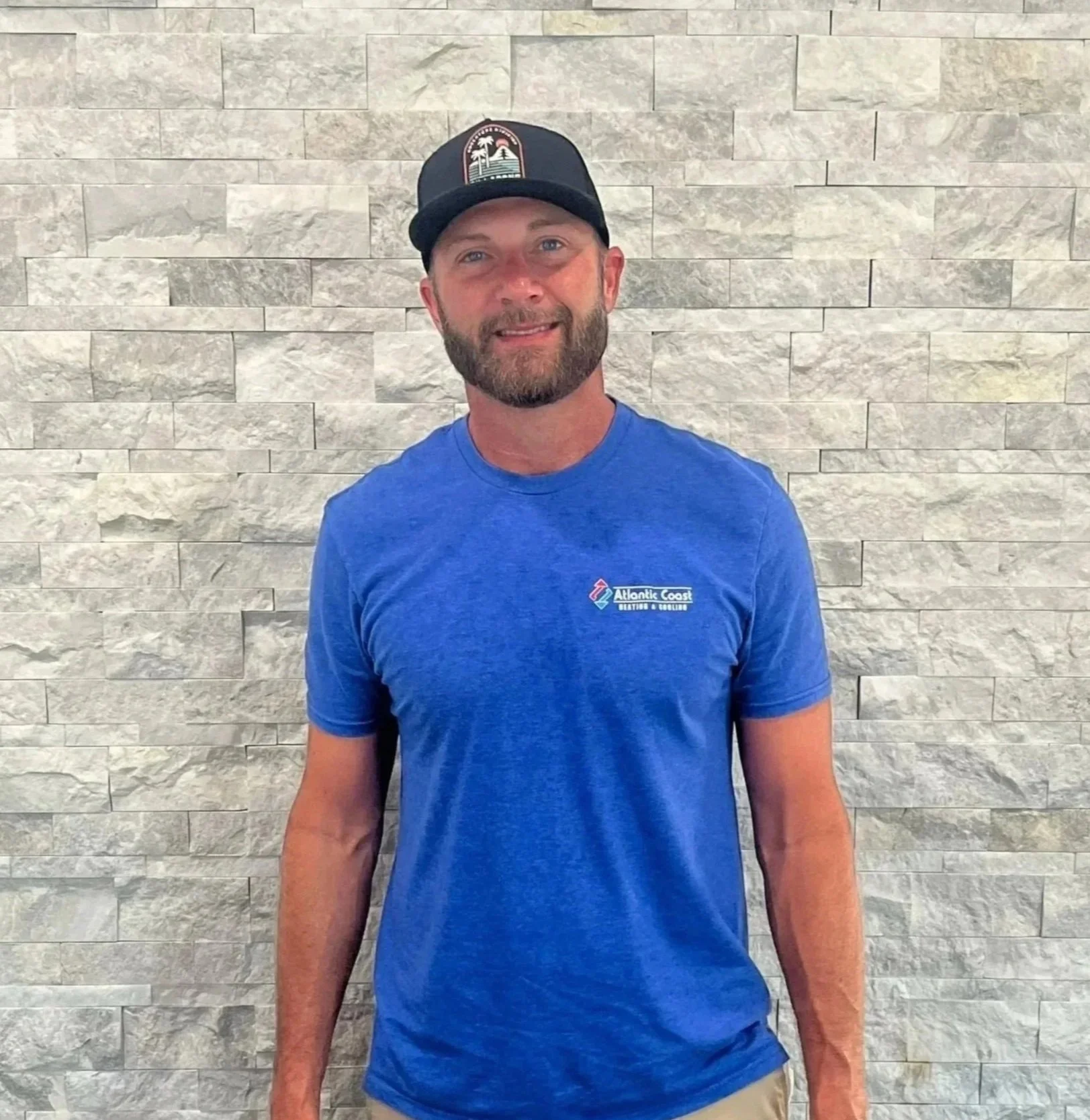 A man with a beard wearing a black cap and a blue t-shirt with 'Atlantic Coast' logo, standing in front of a beige stone wall.