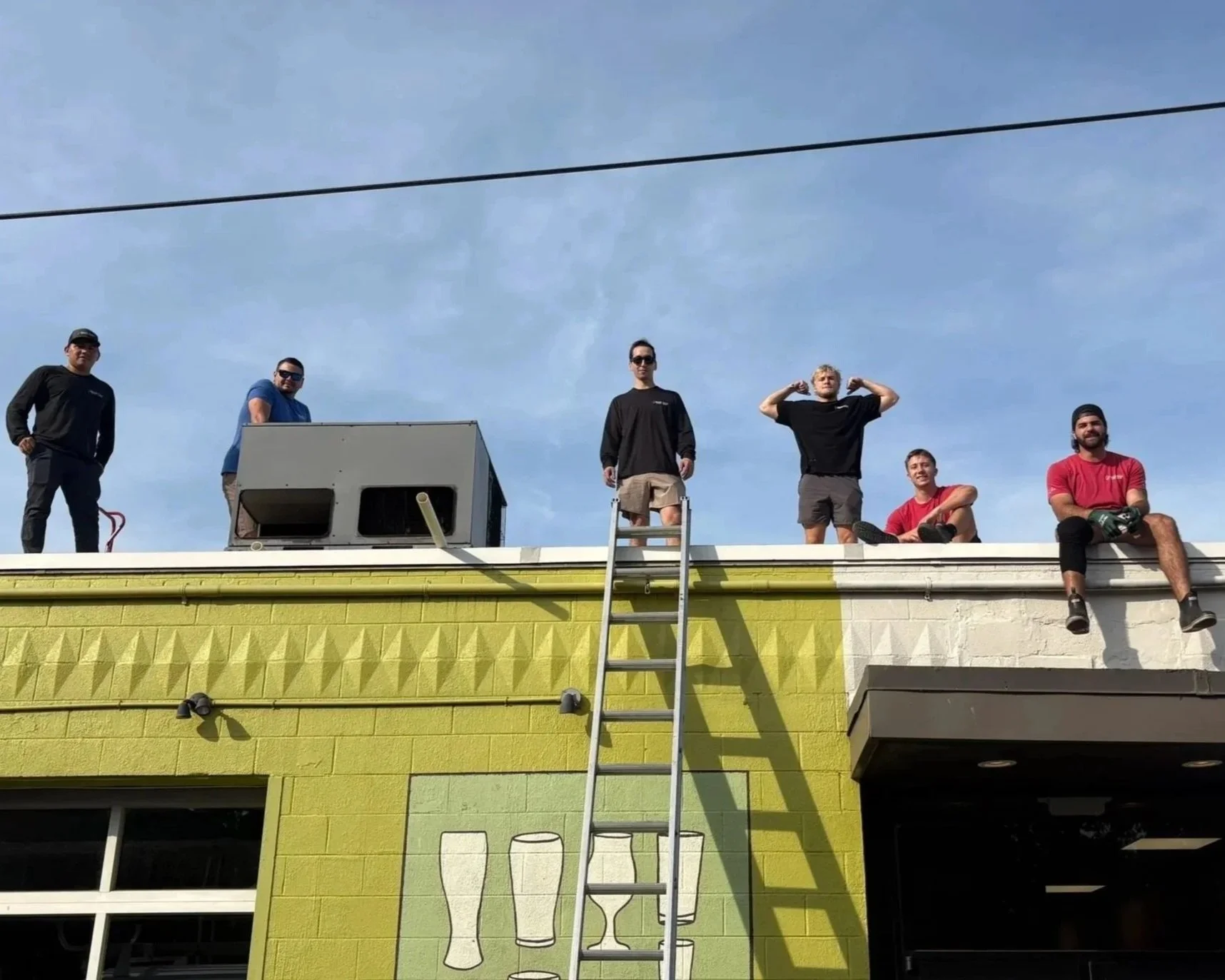 Group of six men on a rooftop, some standing, some sitting, with a ladder leaning against the building, in front of a bright green wall with a mural of glasses, under a clear blue sky.