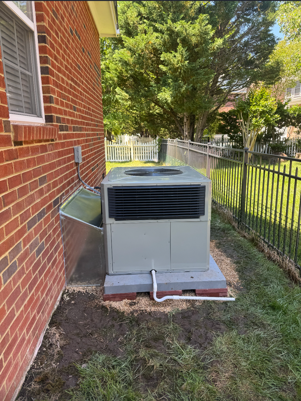 An air conditioning unit outside a house with a brick wall, connected to a vent on the house wall, surrounded by a metal fence and trees in the background.