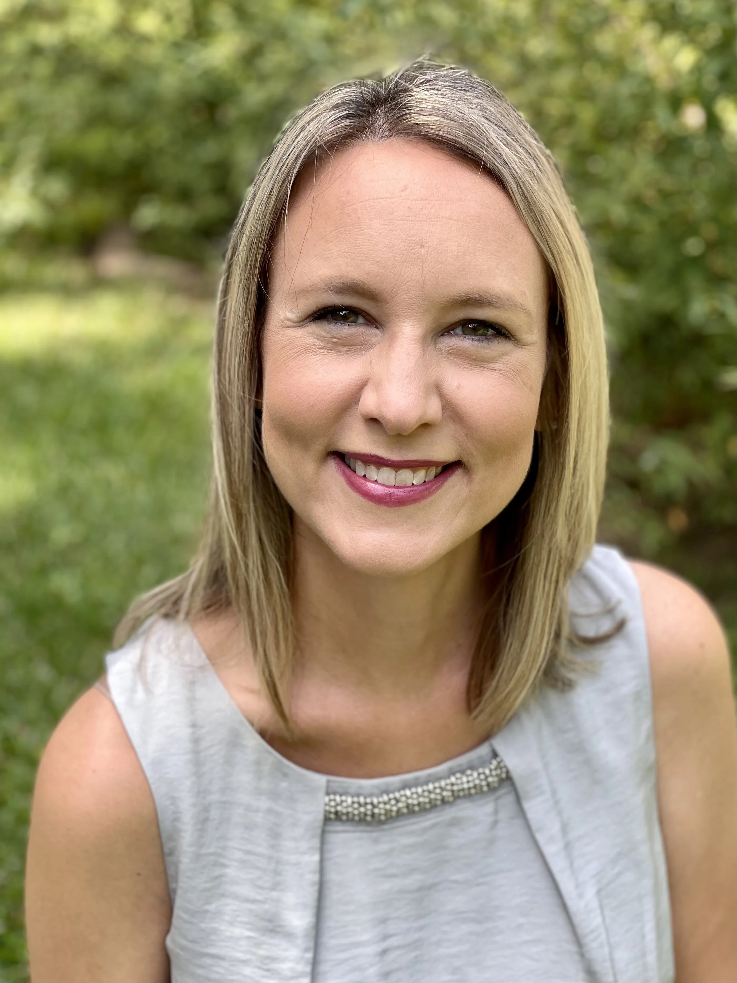 A smiling woman with blonde hair wearing a sleeveless light gray top with a beaded neckline, outdoors with green trees and grass in the background.