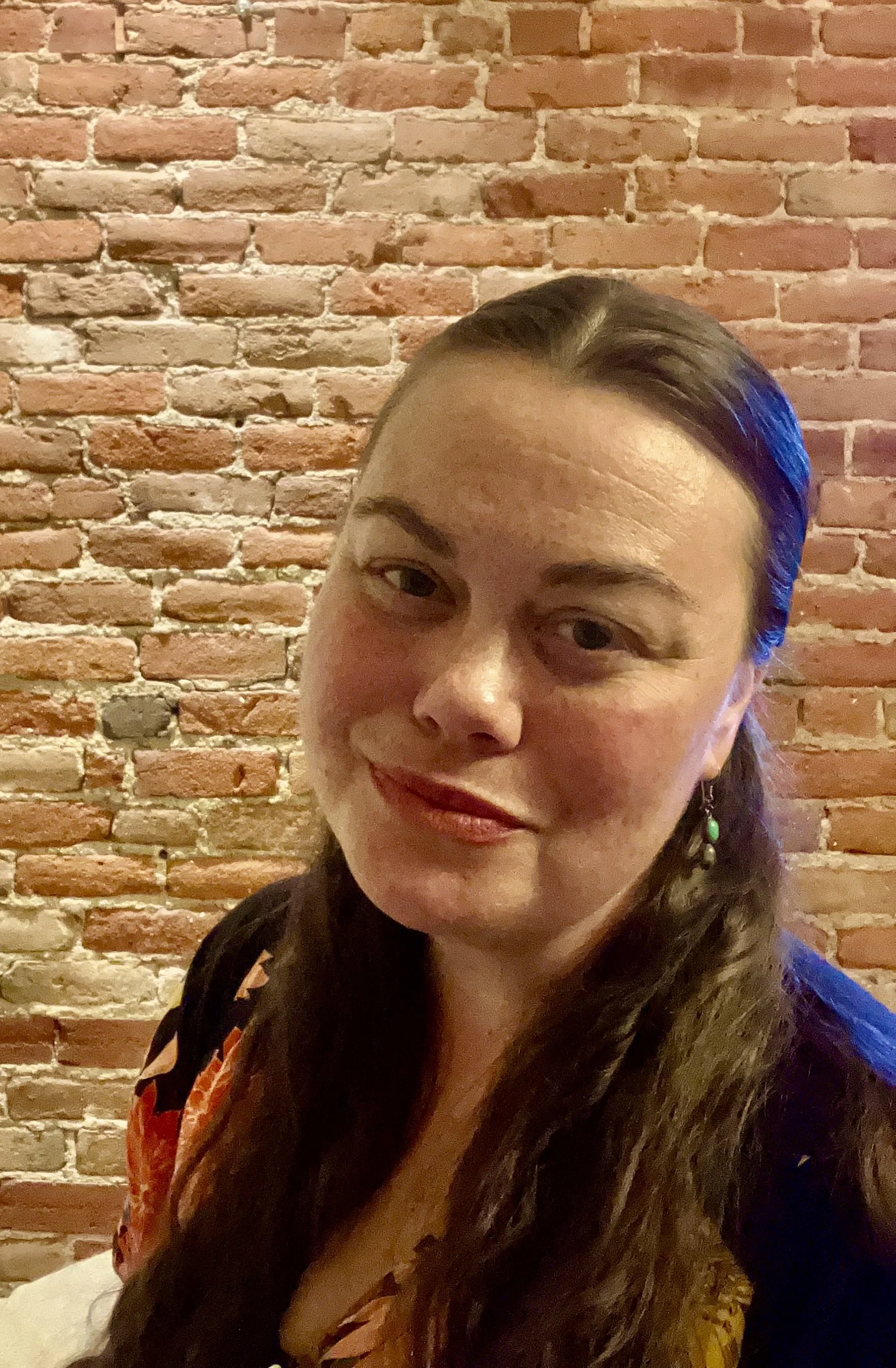 A woman with long dark hair and earrings standing in front of a brick wall, smiling slightly.