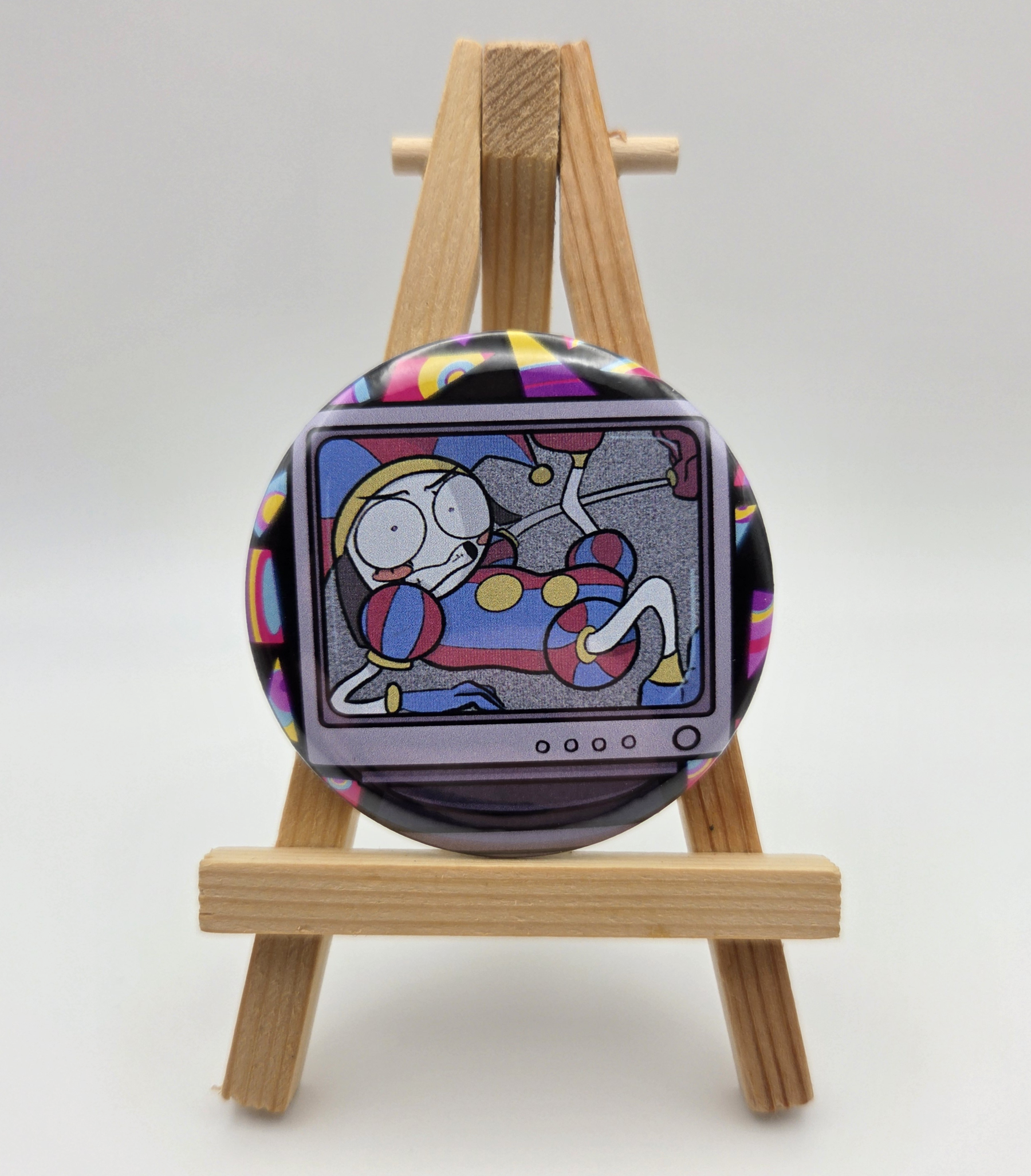 A button featuring POMNI from THE AMAZING DIGITAL CIRCUS resting on a small easel.