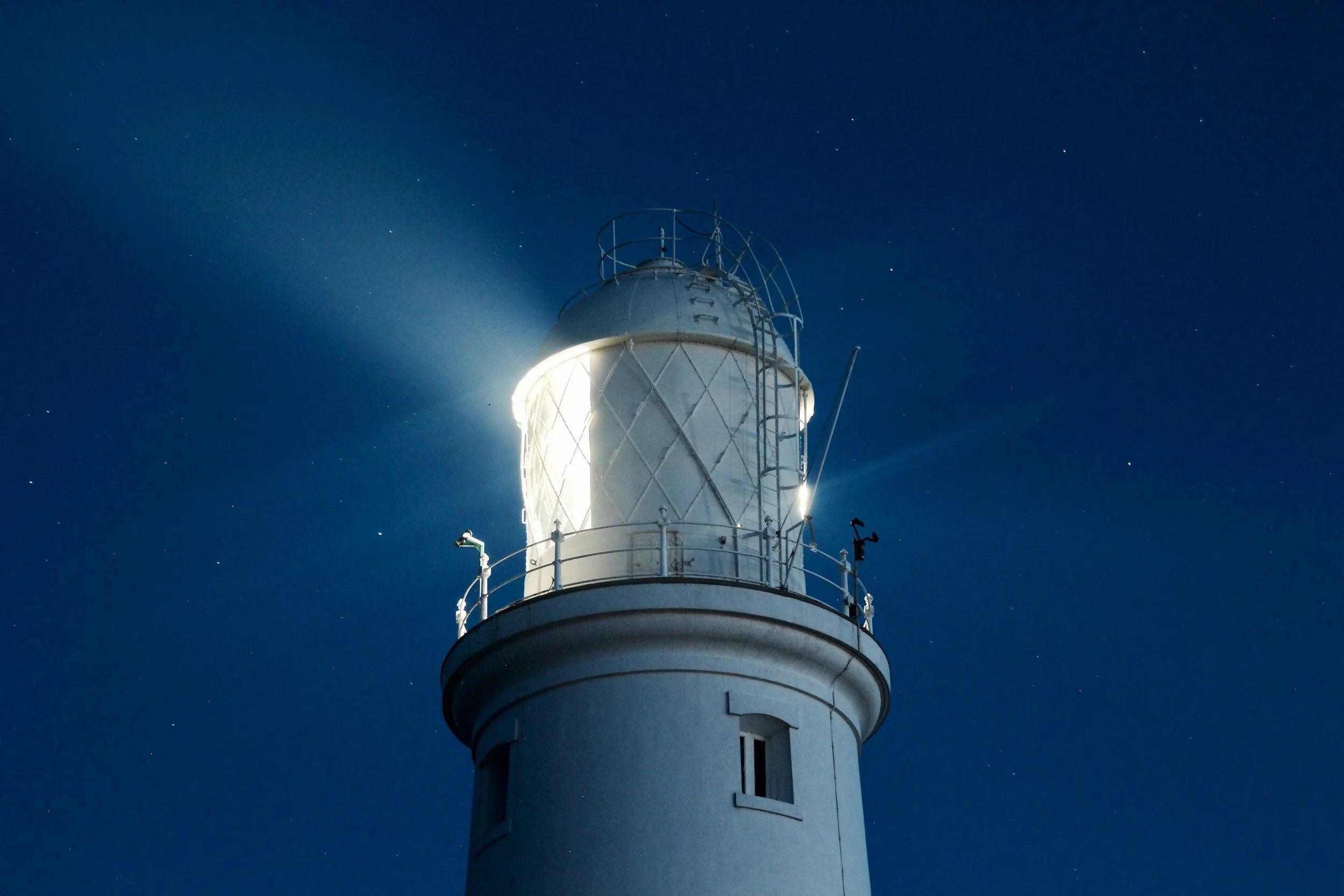 An offshore lighthouse emitting light at night with a clear starry sky in the background.