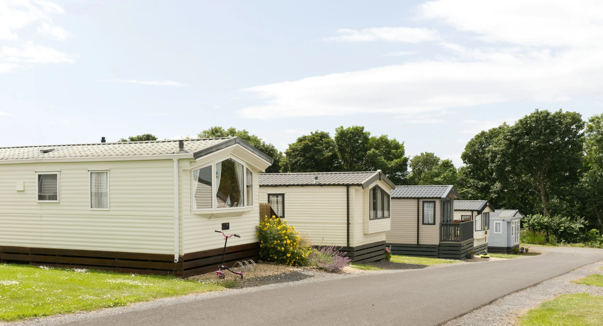A row of white and beige mobile homes with dark gray roofs, situated along a paved street, with green trees in the background under a partly cloudy sky.