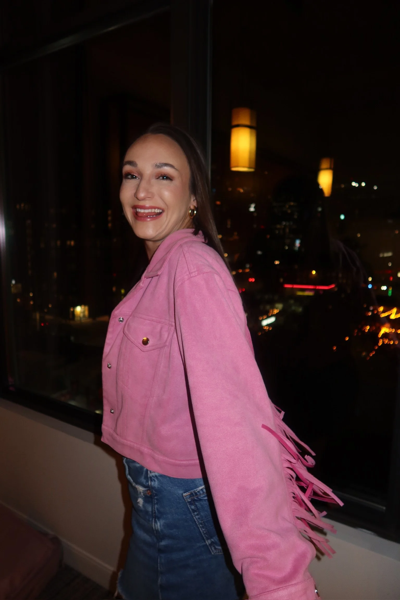 A woman with long brown hair wearing a pink jacket and jeans, smiling and winking at the camera, standing near a large window at night with city lights outside and hanging yellow-orange lamps inside.
