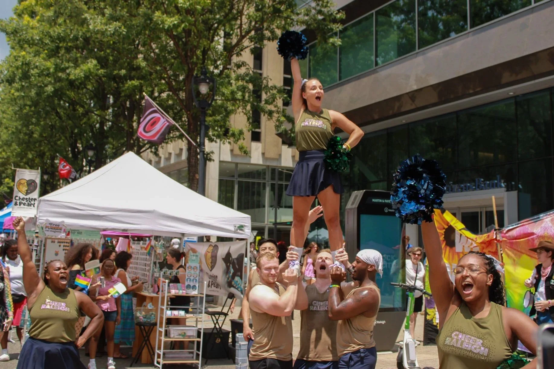 A diverse group of people celebrating at a pride event with colorful flags and banners, performing a cheer stunt with one person lifted high.
