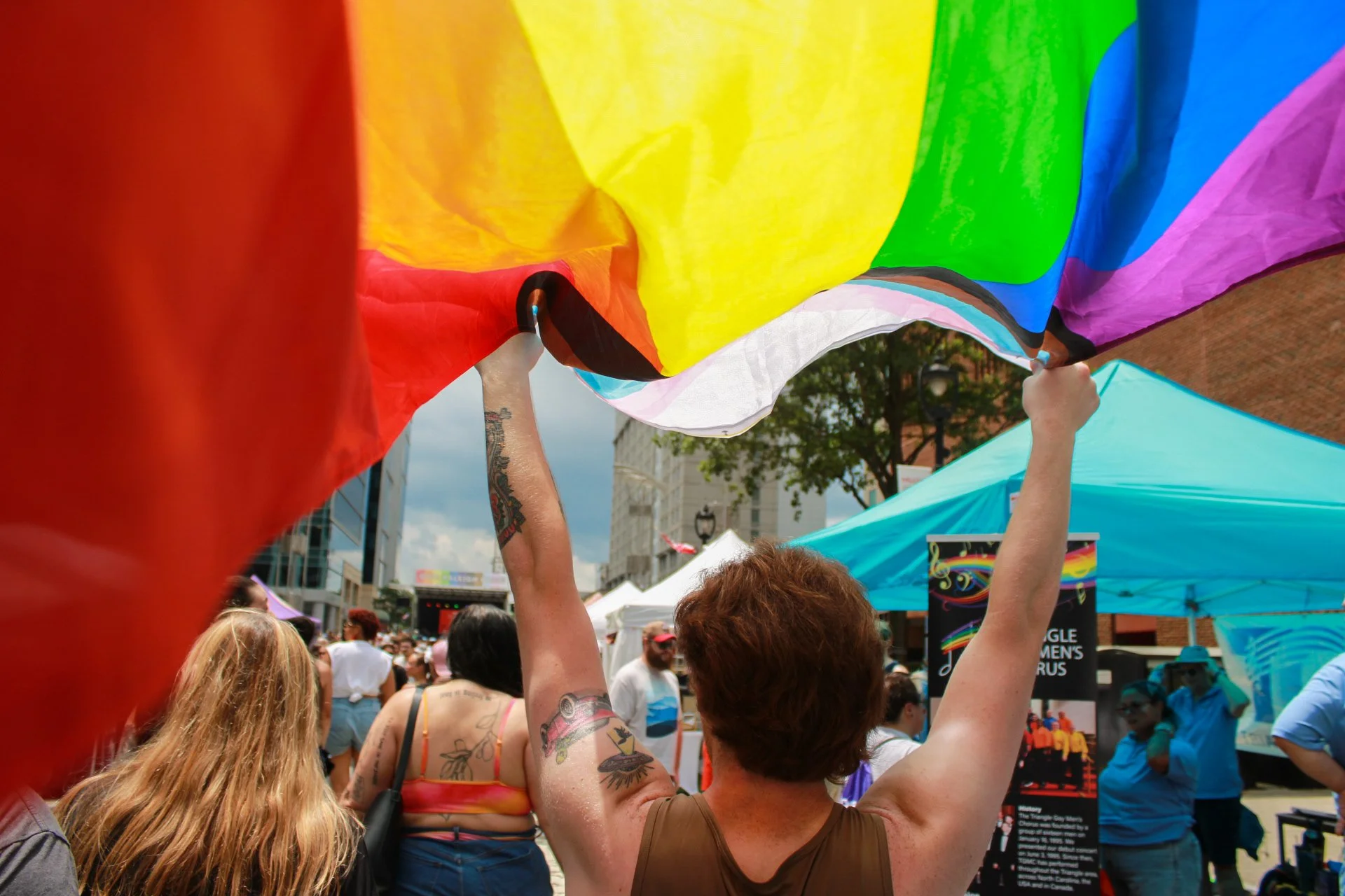 People holding up a rainbow flag during a pride festival on an outdoor city street, with tents and buildings in the background.