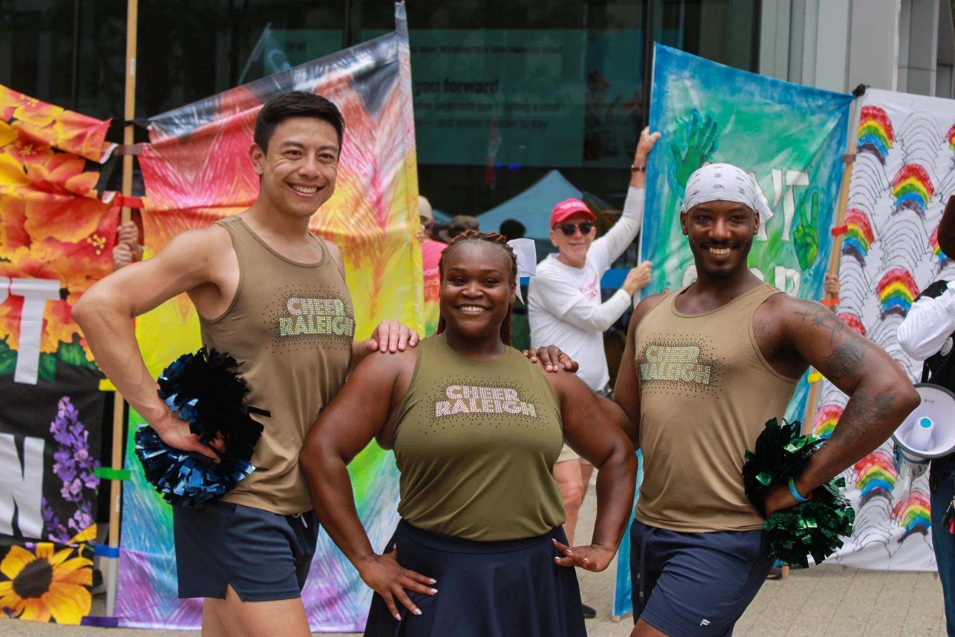 Three cheerleaders posing at an outdoor event, wearing gold tank tops with the words 'CHEER RALEIGH' in rhinestones, smiling for the camera. Behind them are colorful banners and flags, with other participants in the background.