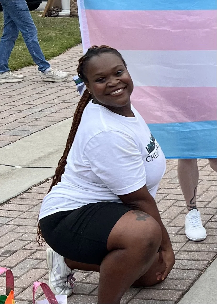 A smiling woman with braided hair in a bun, kneeling on one knee on a brick walkway, wearing a white t-shirt with a logo and black shorts, at an outdoor event with pride flags in the background.