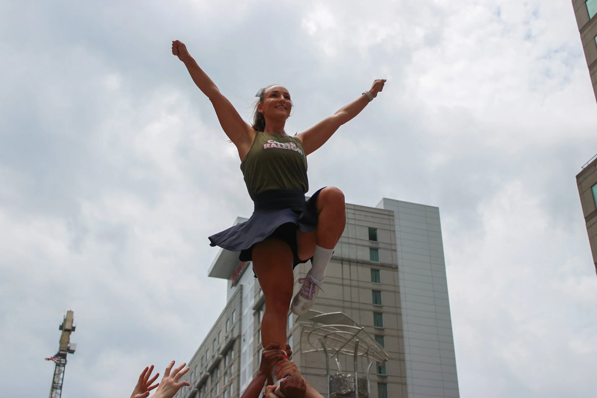 A woman in athletic clothing performing a stunt on a human pyramid during an outdoor event in front of tall buildings with a cloudy sky.