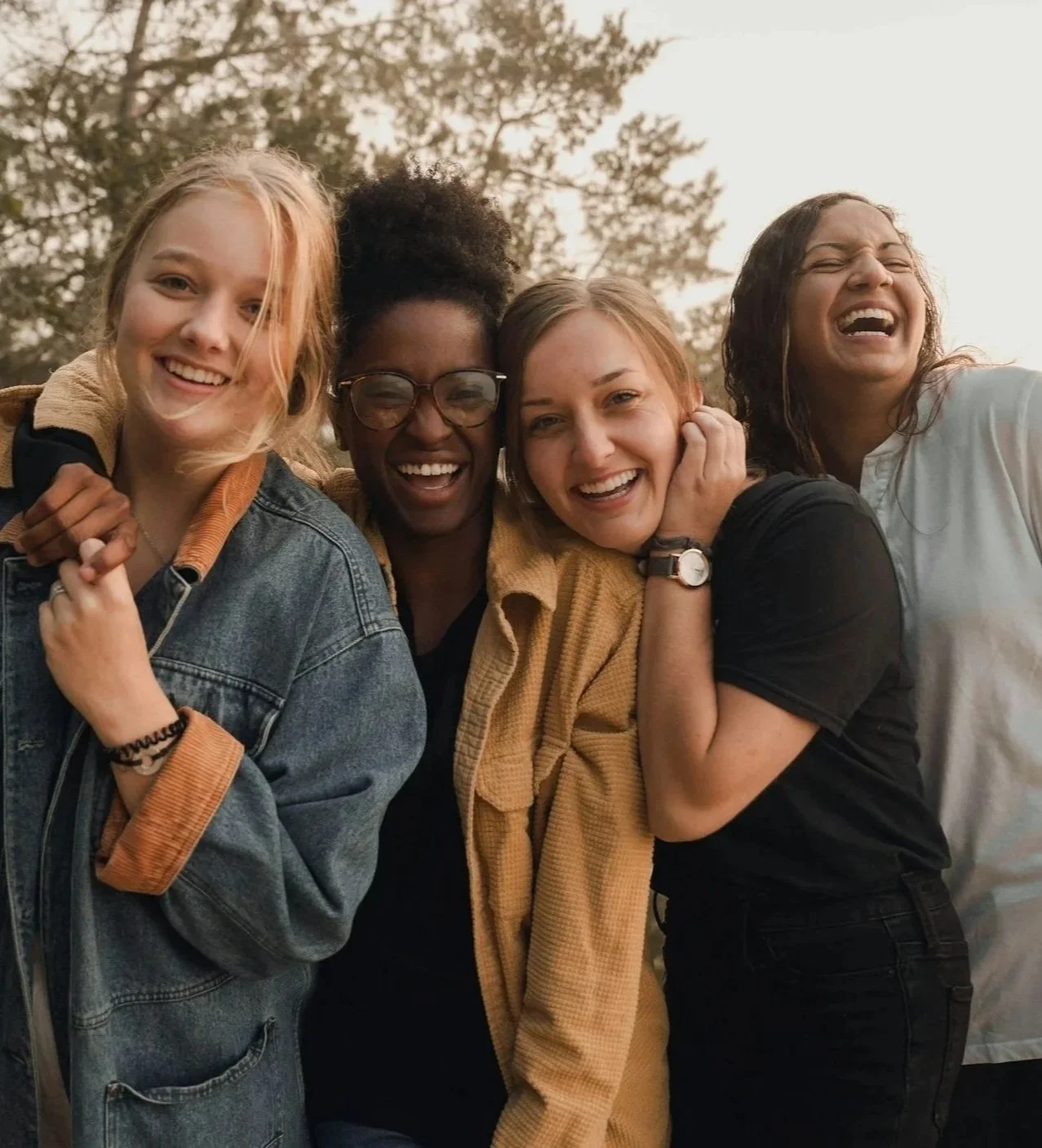 Four women smiling and hugging each other outdoors in warm clothing and casual attire.