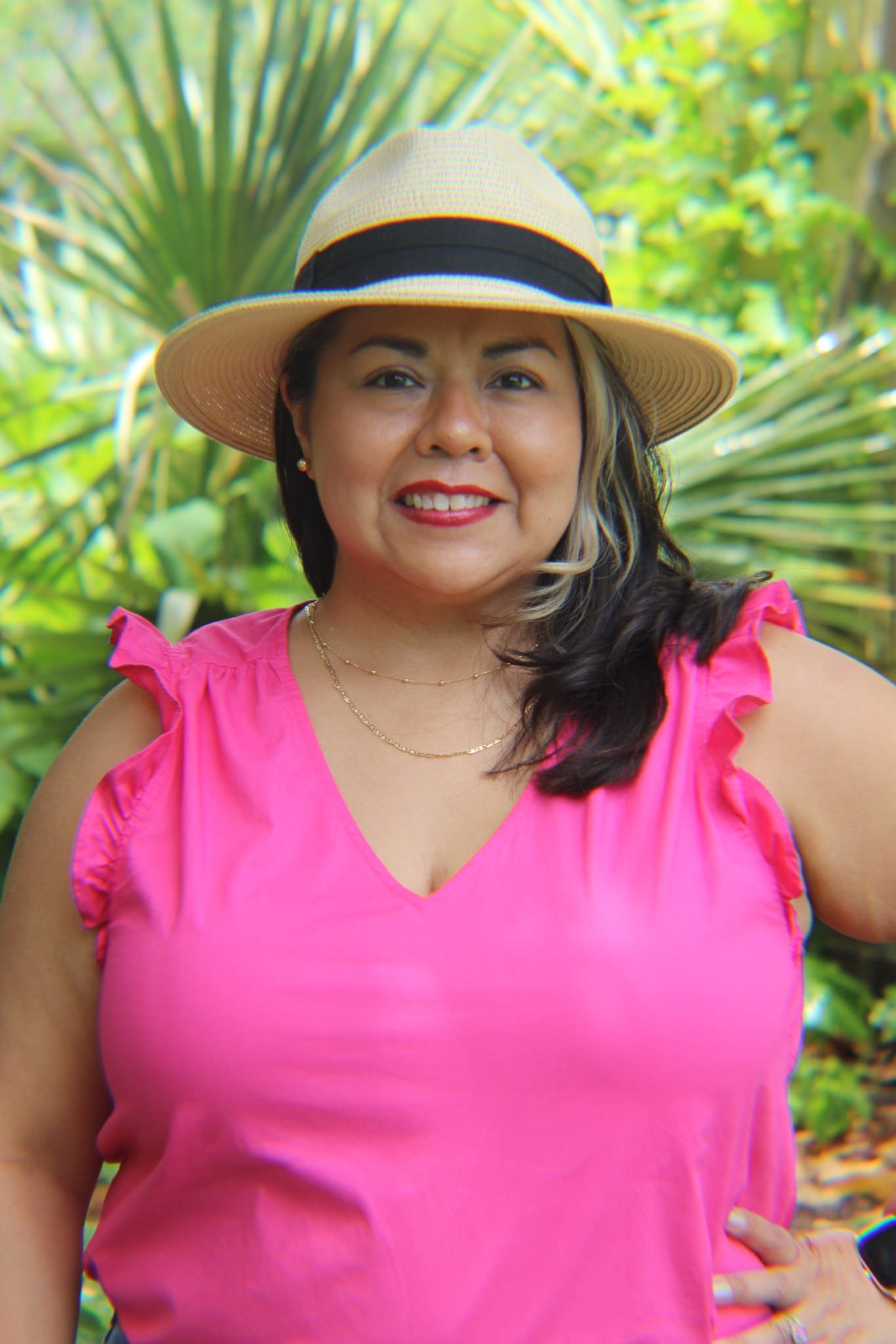 A woman smiling outdoors wearing a pink top and a wide-brimmed straw hat with a black band, standing in front of lush green tropical plants.