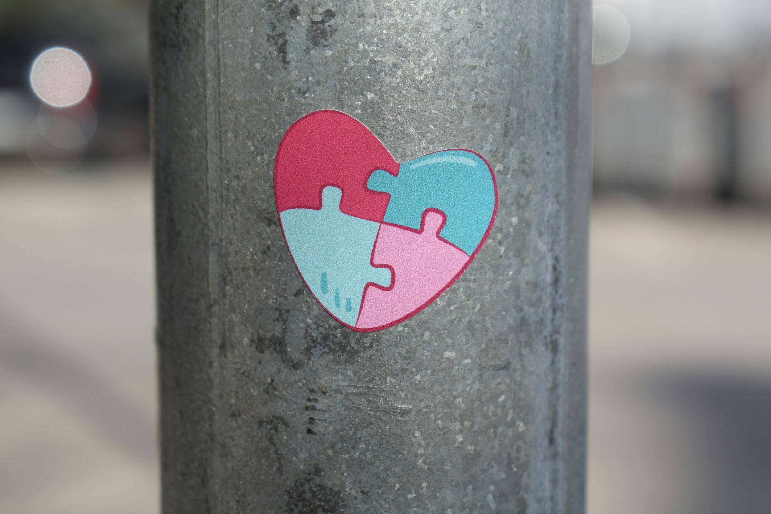 A sticker of a heart-shaped puzzle in pink and blue colors is stuck on a gray concrete surface, with blurred city lights in the background.