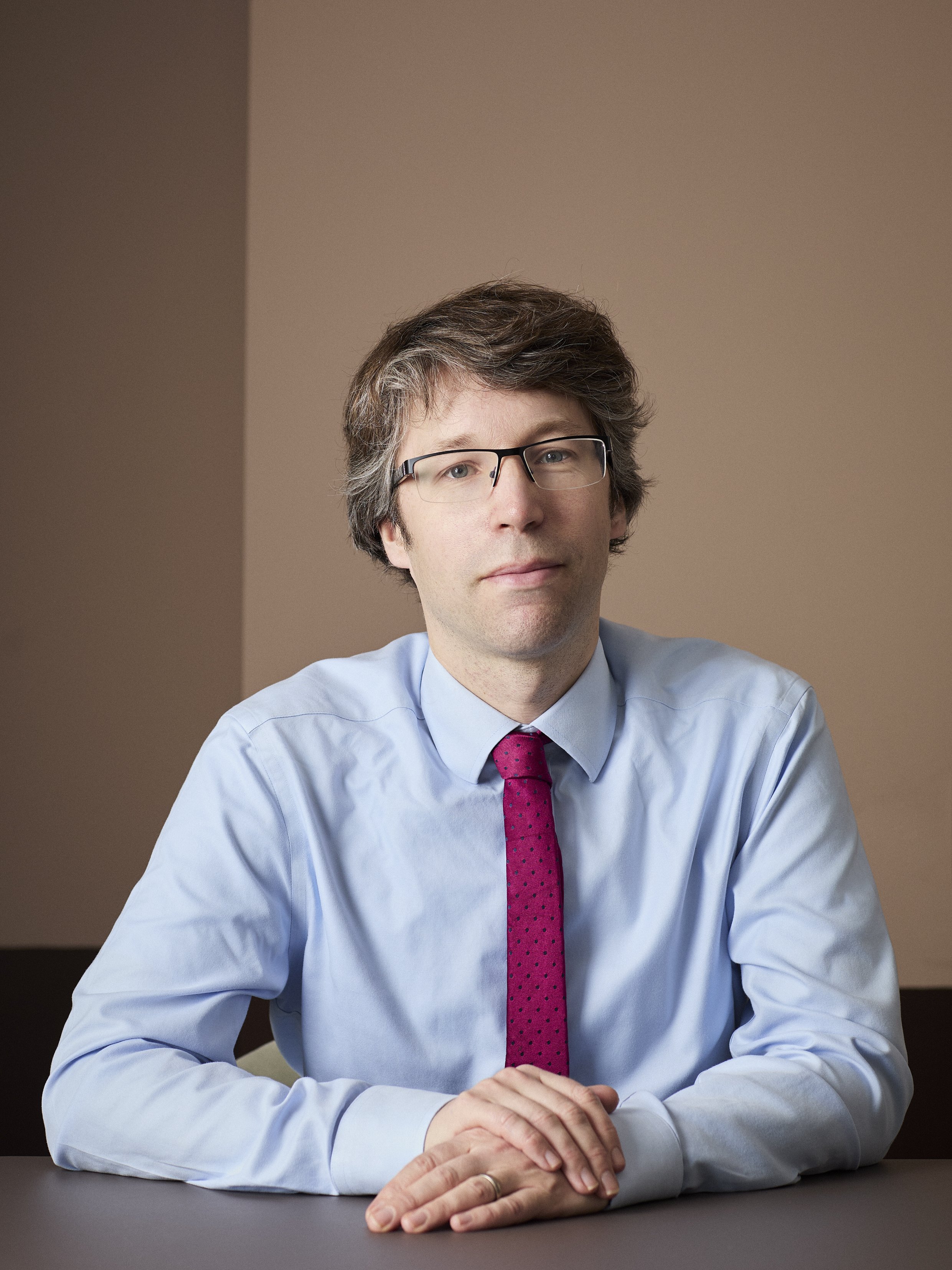 A man with glasses wearing a light blue shirt and a pink tie, sitting at a table with hands folded, against a beige wall.