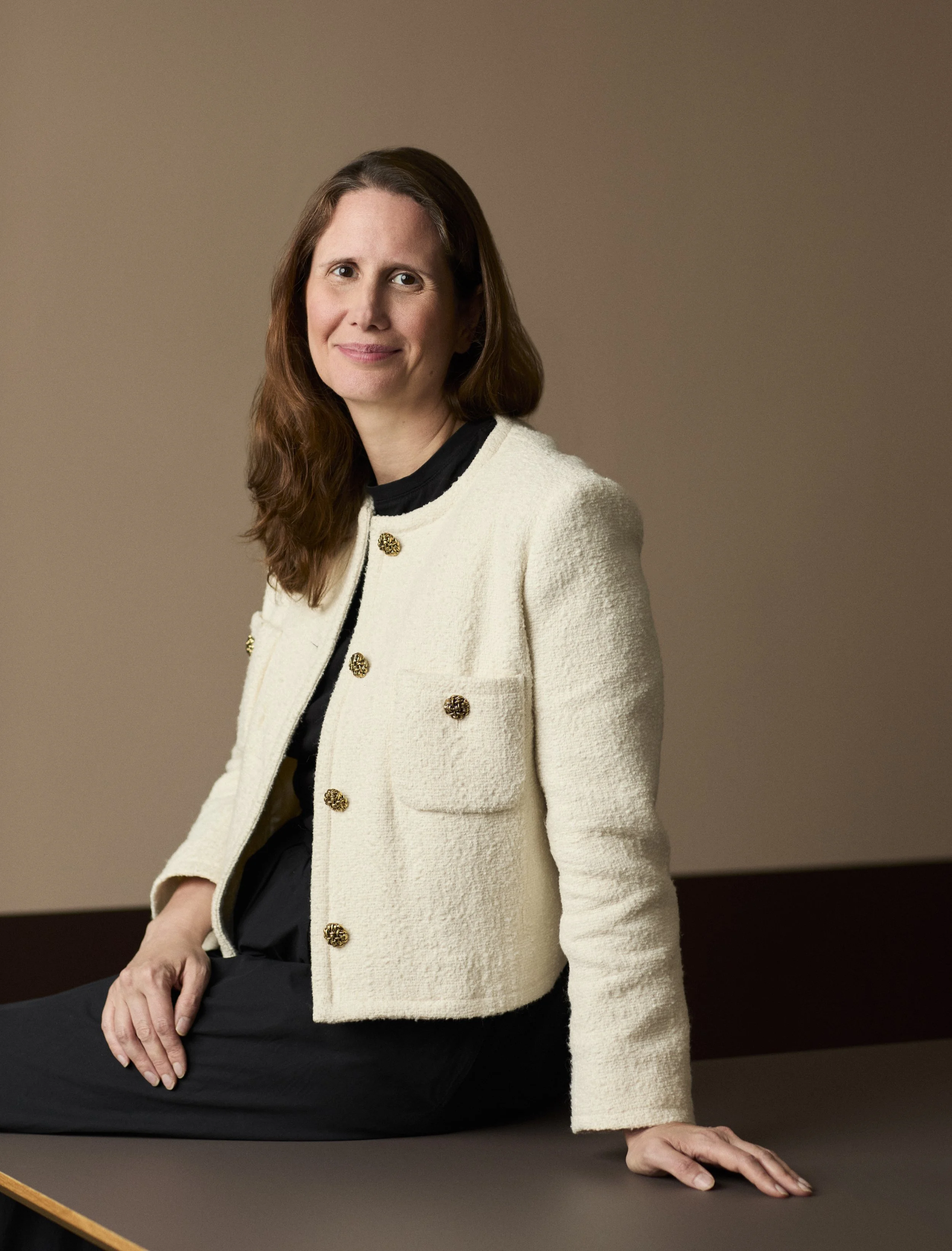 A woman with brown hair wearing a cream-colored coat with gold buttons, sitting at a table with her hand resting on it, against a plain brown background.
