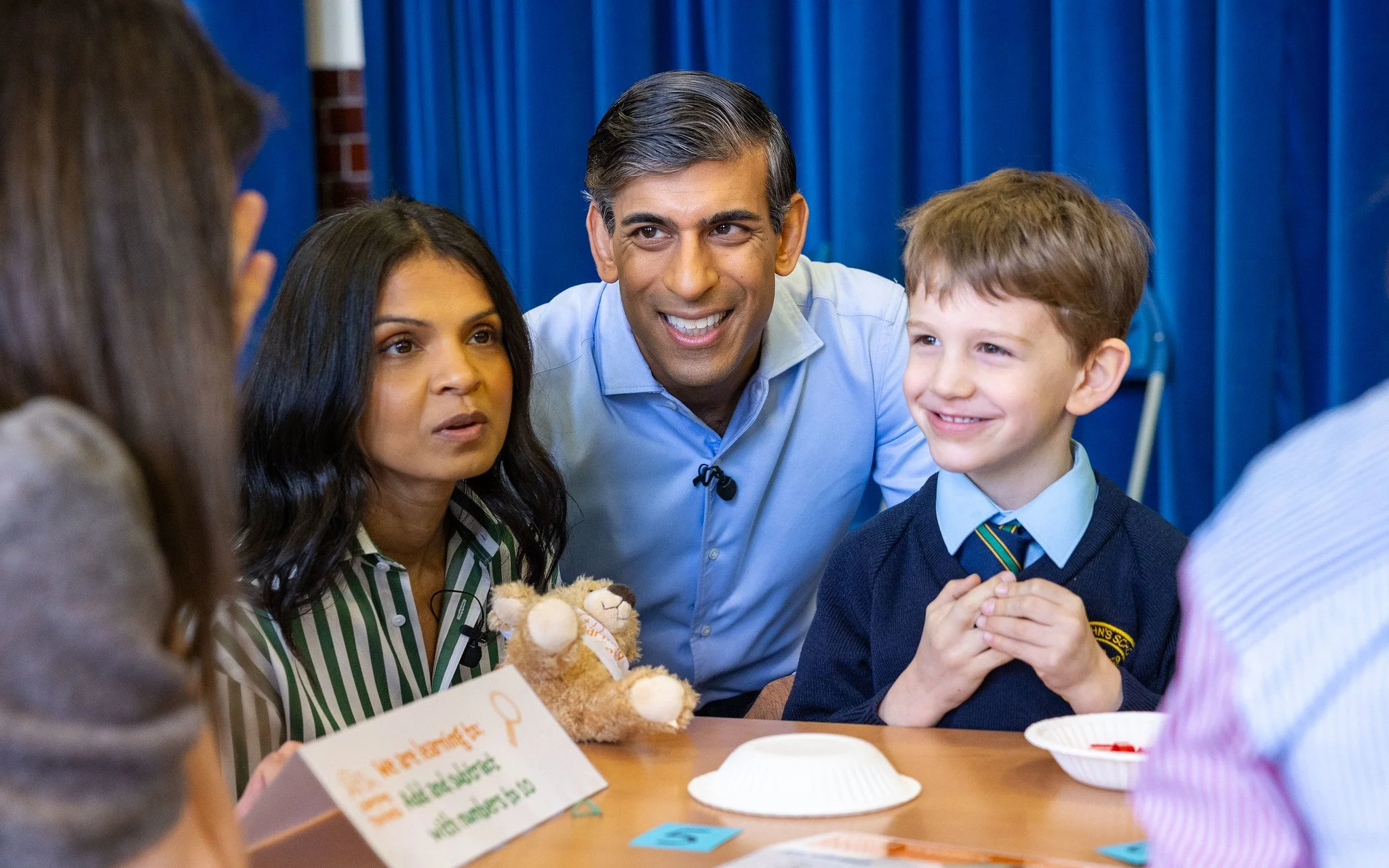 A diverse group of people, including a man, a woman, and a boy in a school uniform, engaged in conversation around a table with food, blue curtains in the background.