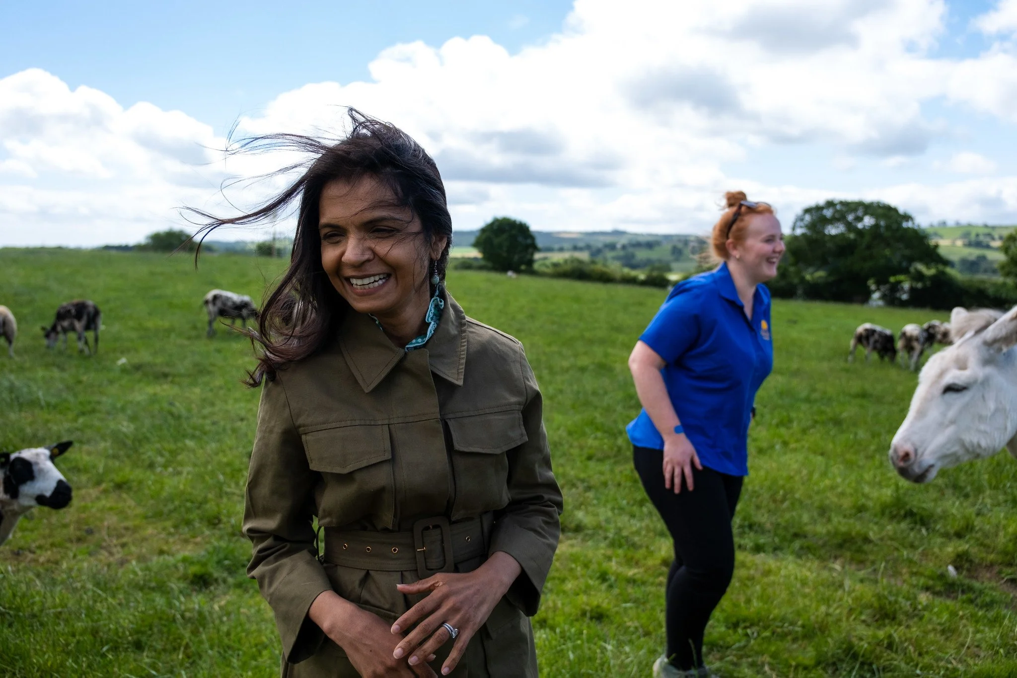 Two women standing in a green field with grazing cows. The woman in the foreground has dark hair, is smiling, and is wearing a brown jacket. The woman in the background has red hair, is wearing a blue shirt, and is laughing. The sky is partly cloudy 