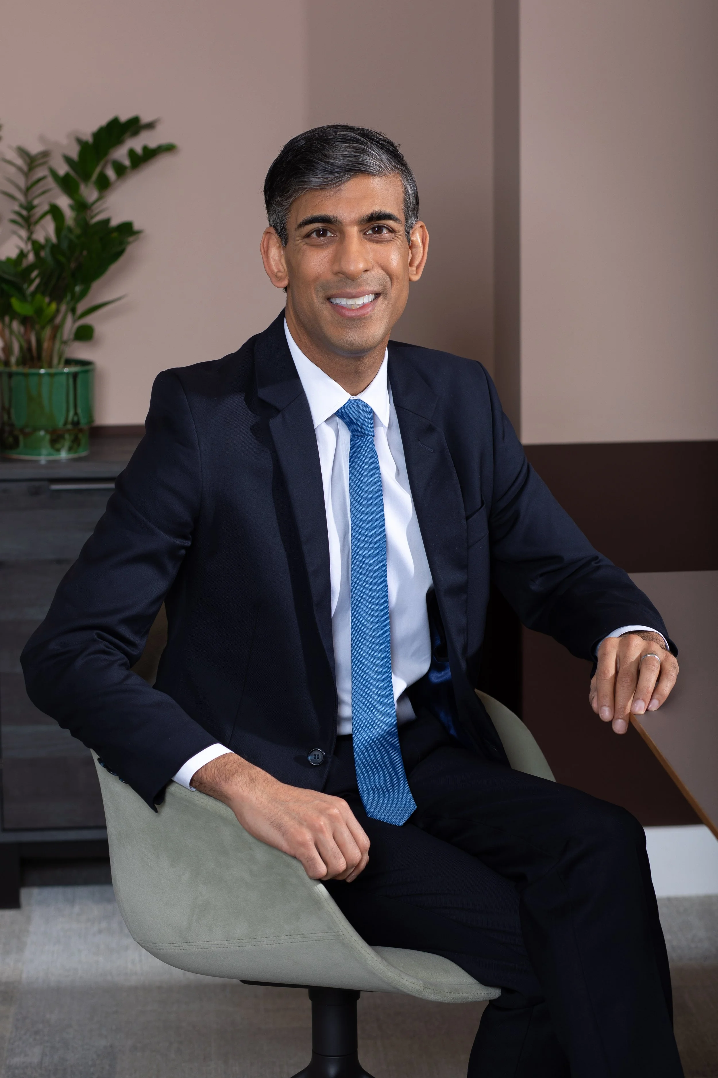 A man in a navy suit, white dress shirt, and blue tie sitting in a modern office chair, smiling at the camera. There is a potted plant on a desk in the background and the office has beige and brown walls.