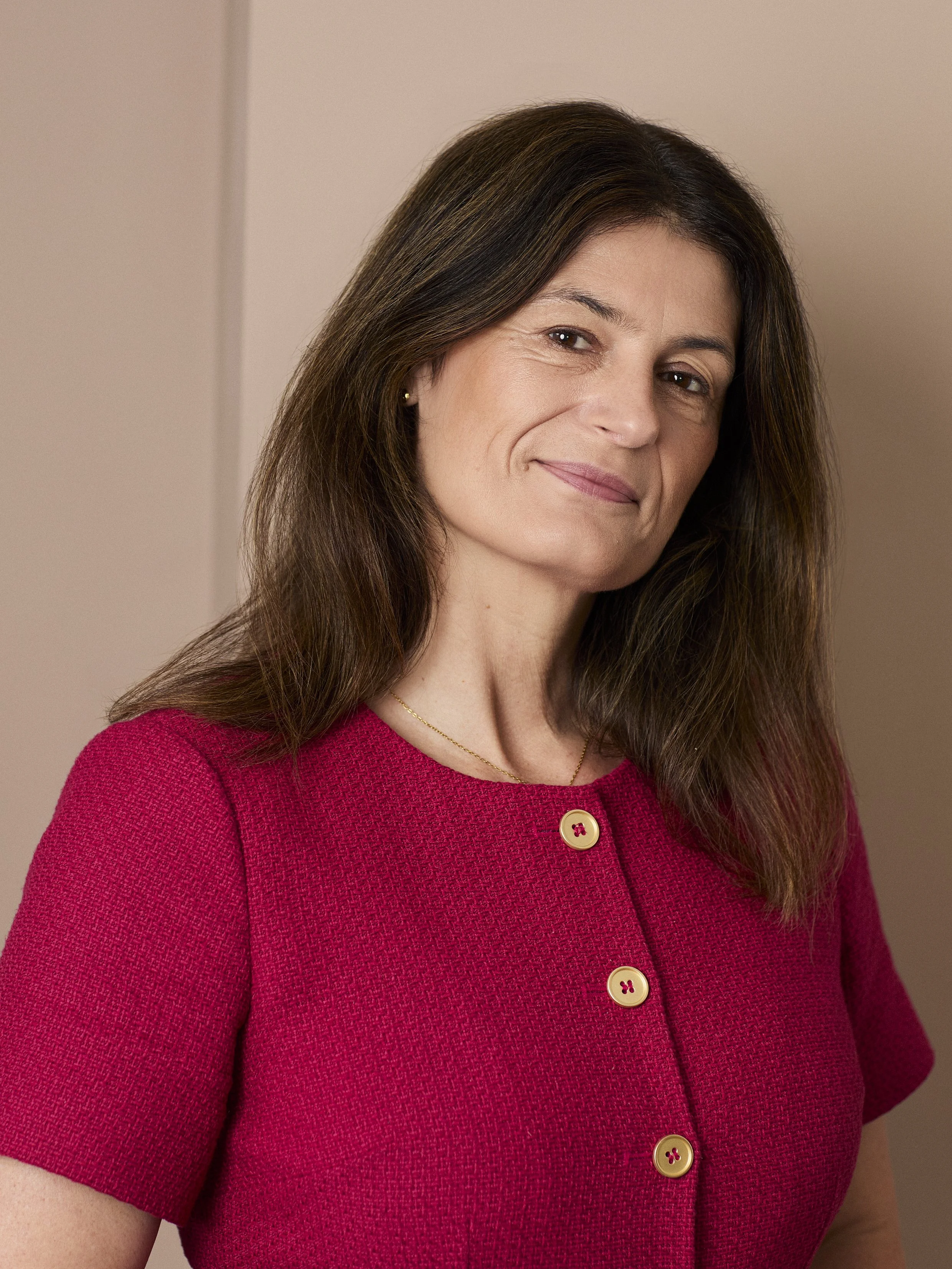 A woman with long brown hair, wearing a red textured top with gold buttons and a delicate gold necklace, smiling softly in front of a plain beige background.