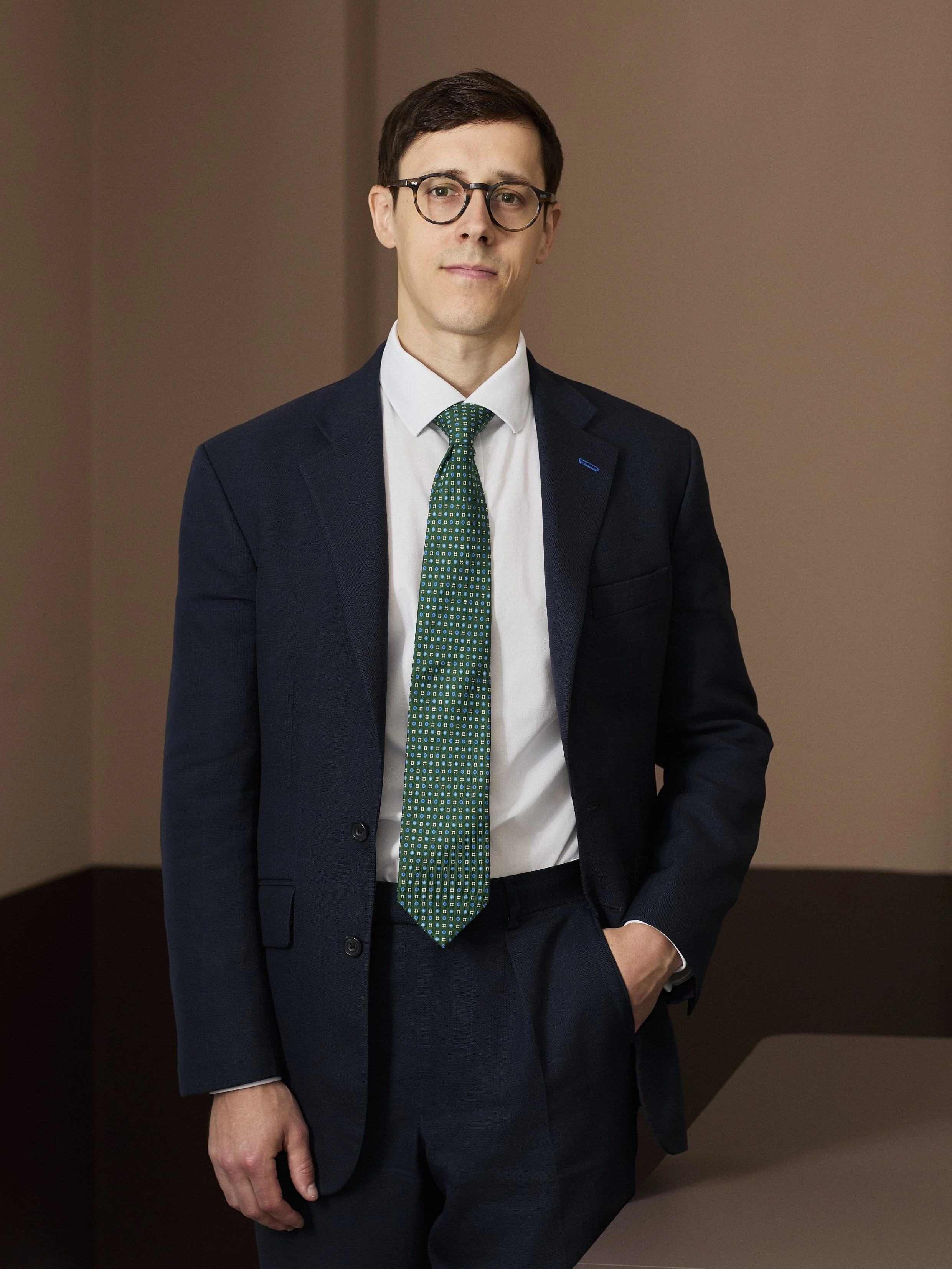 A young man in a dark suit, white shirt, and patterned green tie standing in front of a plain wall.
