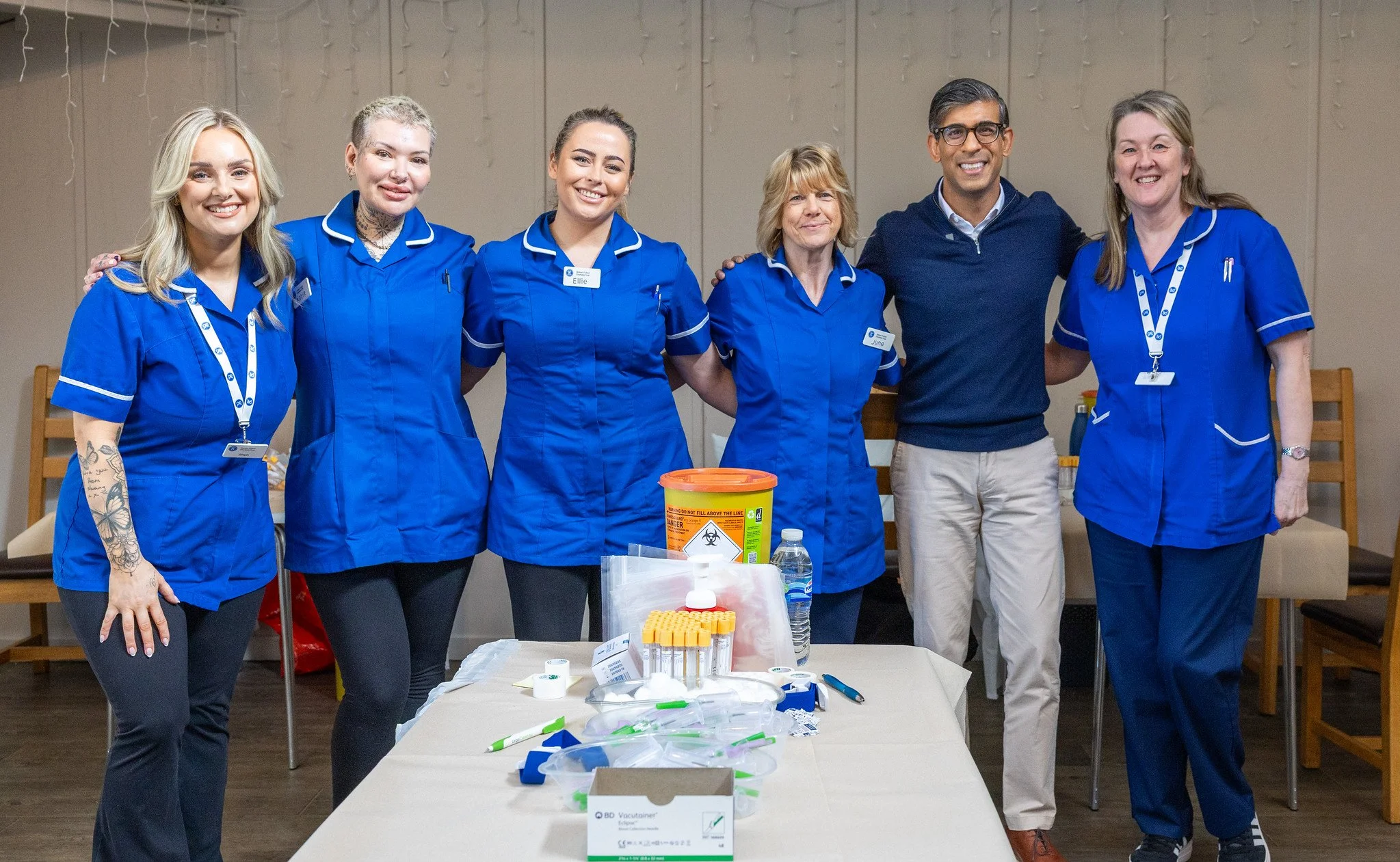 Group of six people standing behind a table filled with medical supplies, smiling at the camera. The group includes five women and one man, most wearing blue uniforms, inside a room with beige walls.