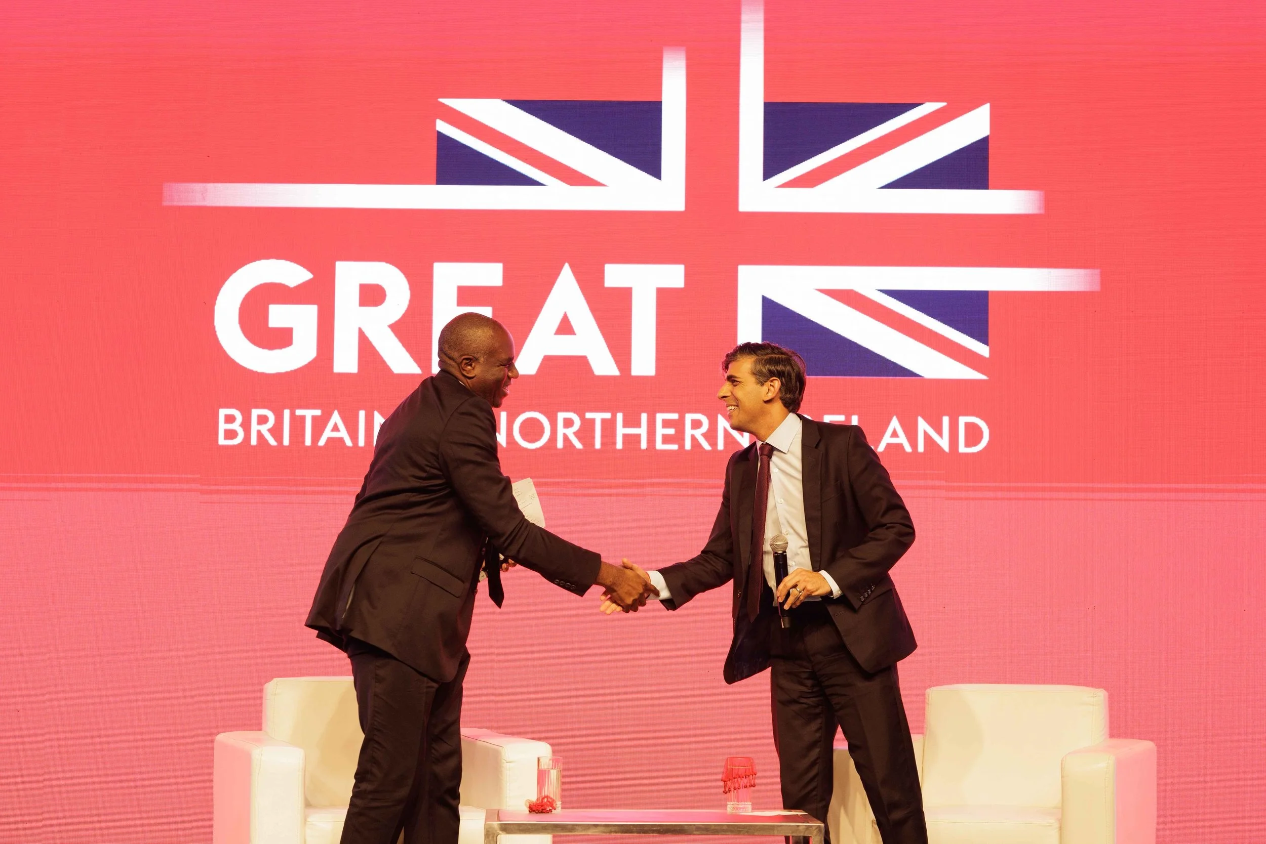 Two men shaking hands on stage at a conference, with a large screen behind them displaying a UK flag and the words "GREAT Britain, Northern Ireland."